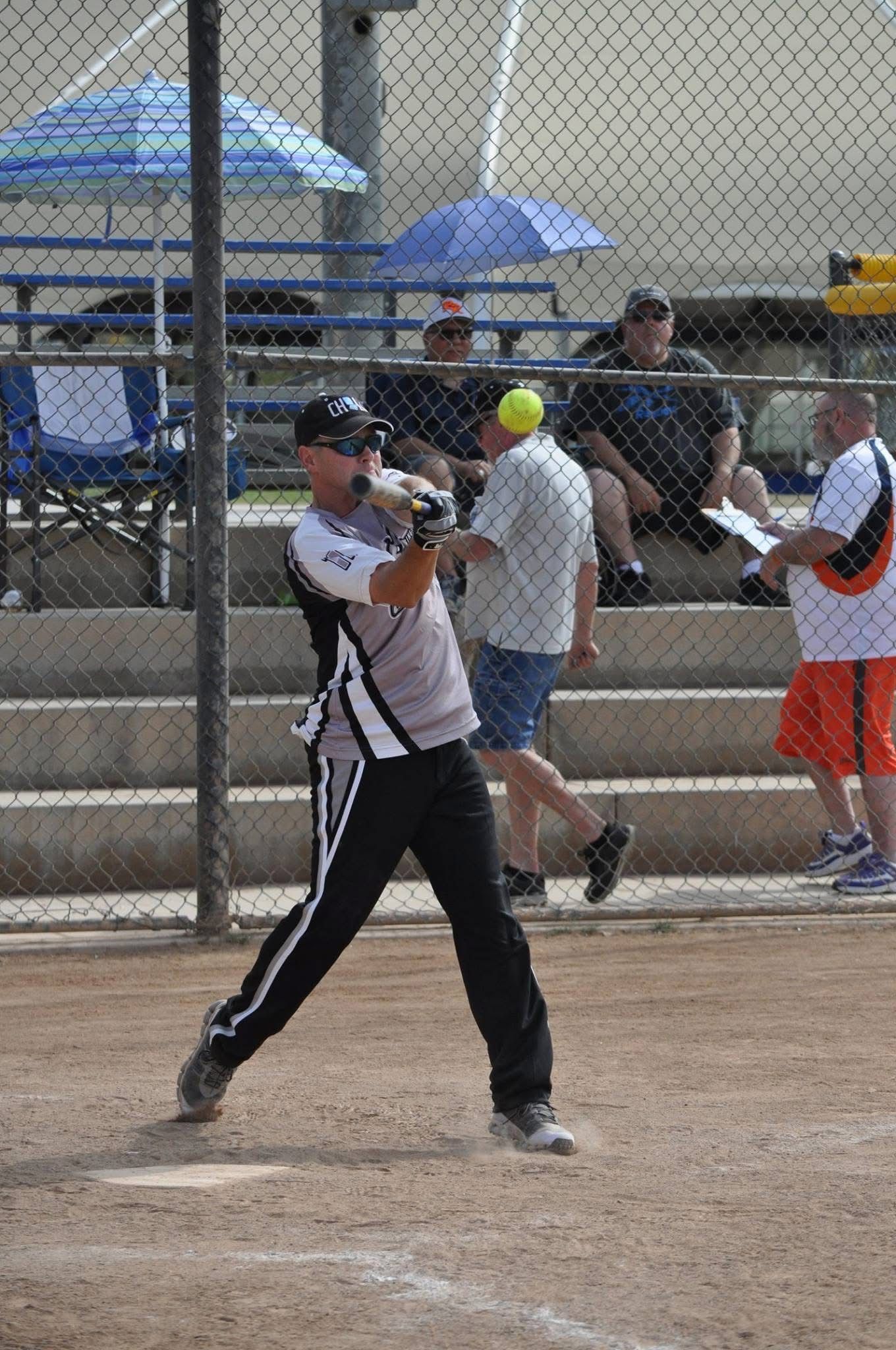 Baseball batter in black and white uniform swinging at a pitch on a dirt field, with spectators behind a fence.