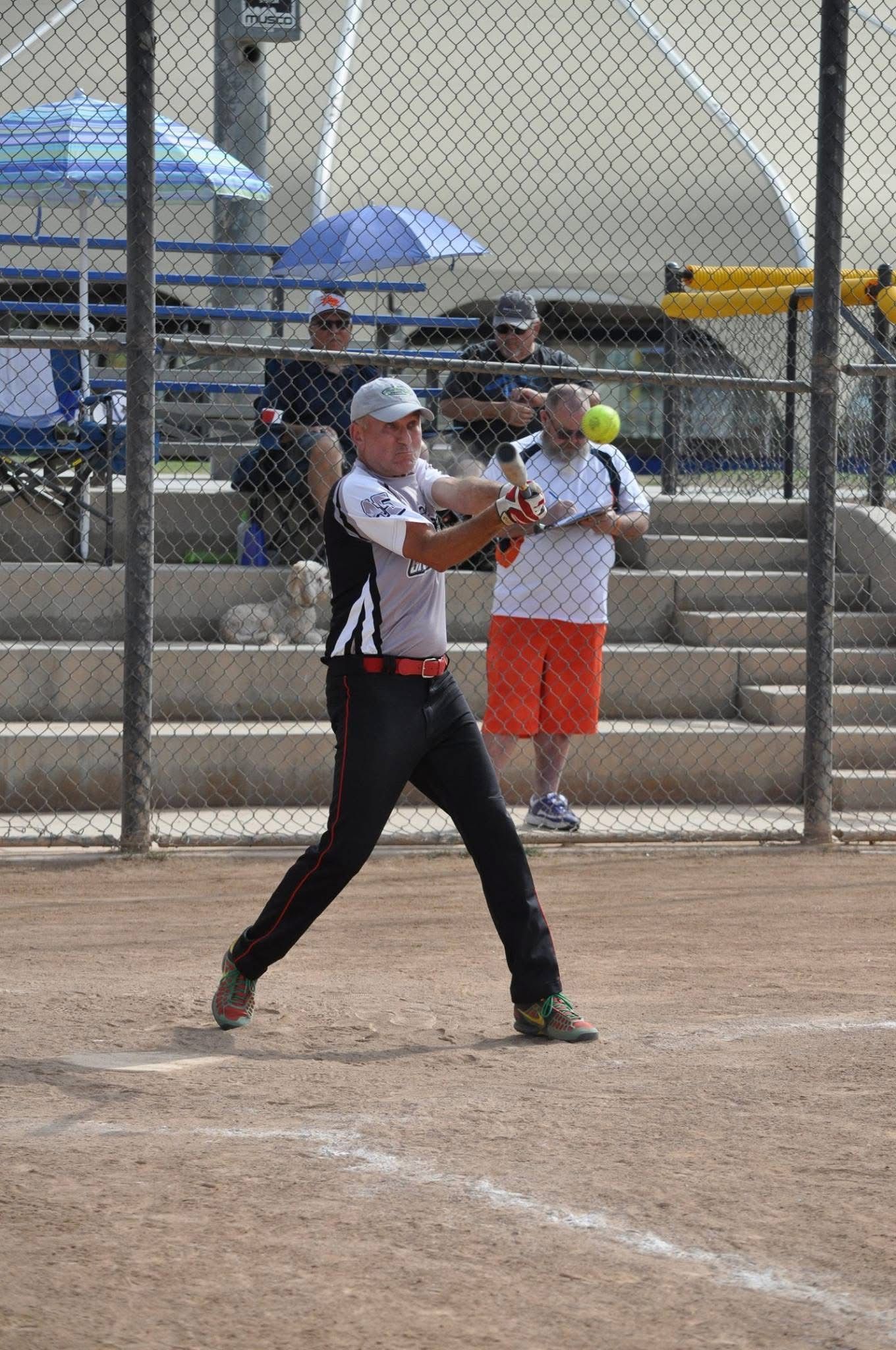 Softball pitcher in black-and-white uniform mid-throw on a dirt field, with bleachers and umbrellas behind her