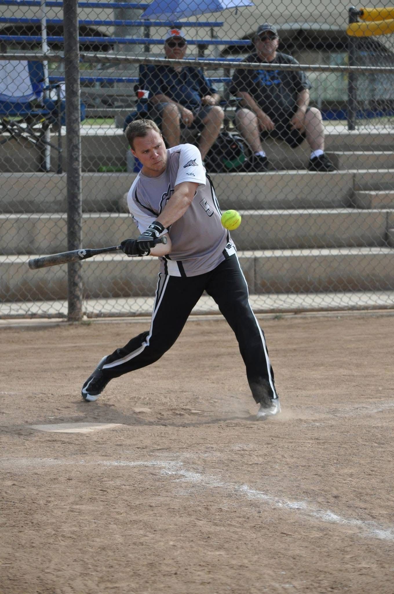 Softball player swinging a bat on a dirt field, with bleachers and spectators in the background.