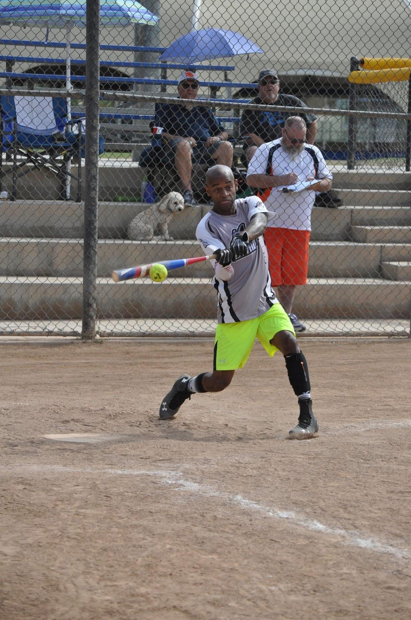 Youth baseball player in black and neon shorts swings a bat on a dirt field, with bleachers behind.