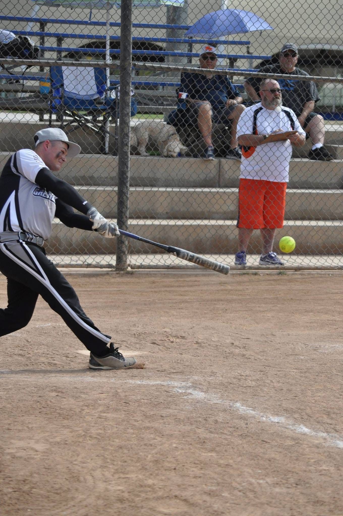 Softball pitcher mid-throw on a dirt field, with spectators behind a chain-link fence.