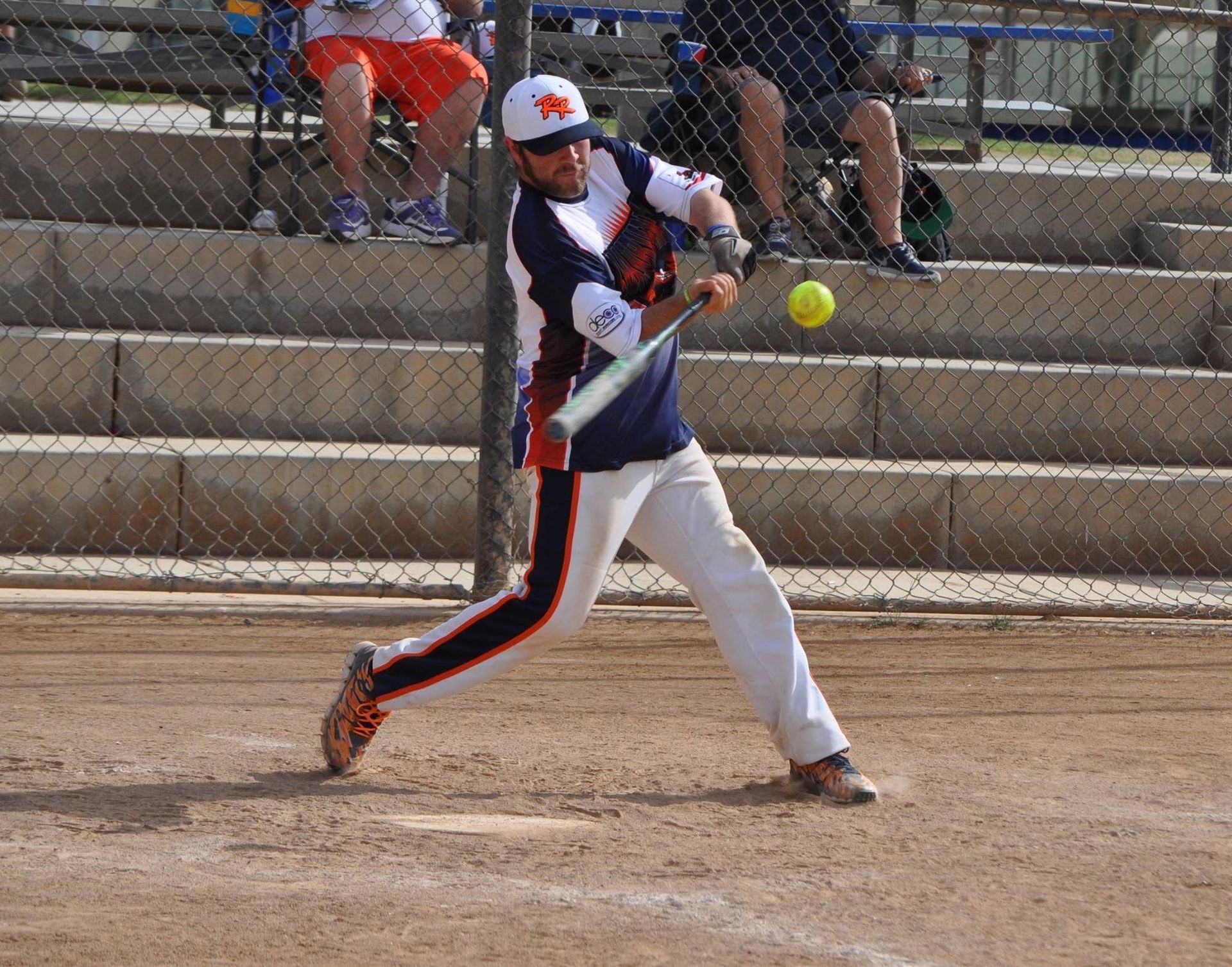 Softball player in navy and white swings at a pitch on a dirt field, with bleachers and spectators behind.