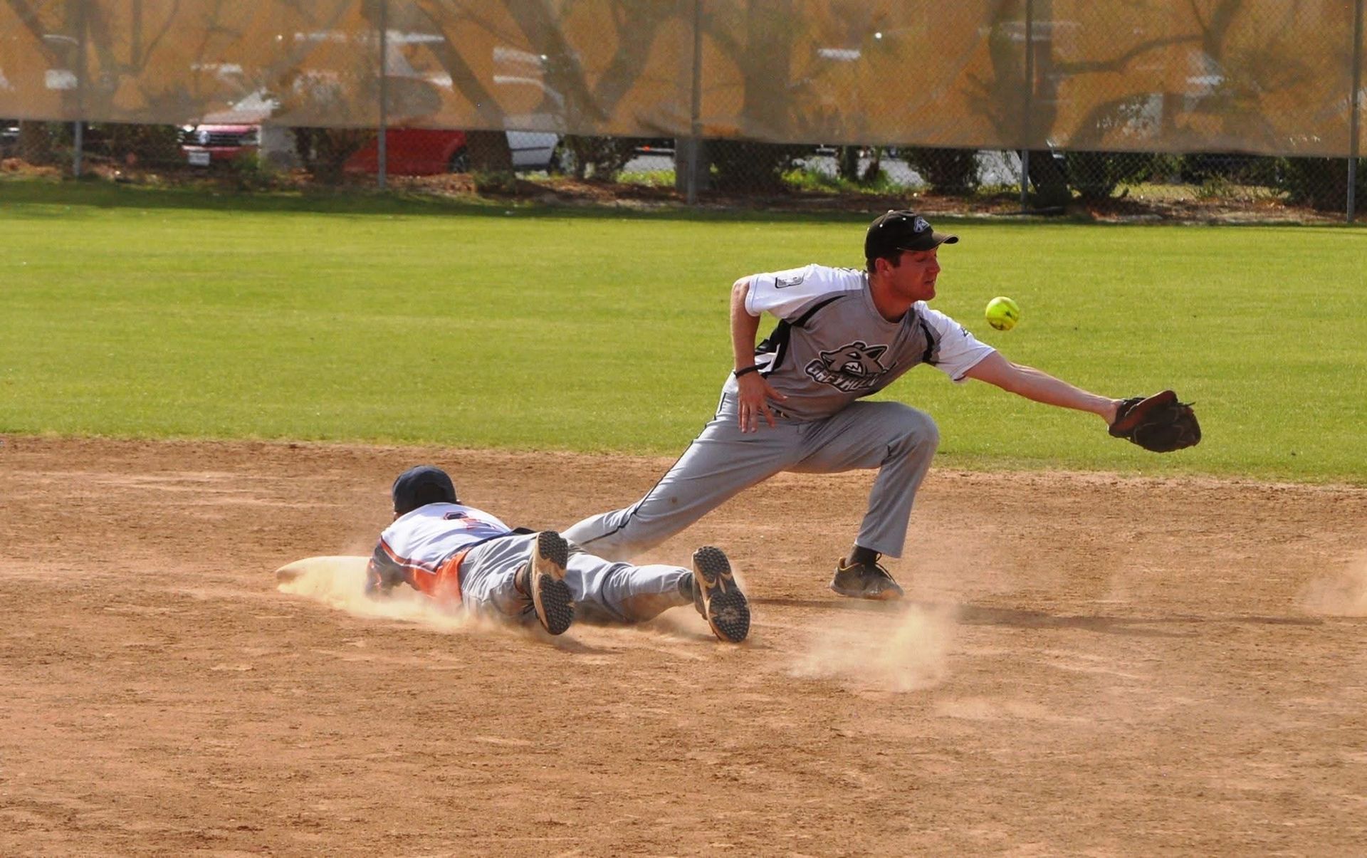 Baseball player in gray lunges to tag a sliding runner at second base on a dusty infield.