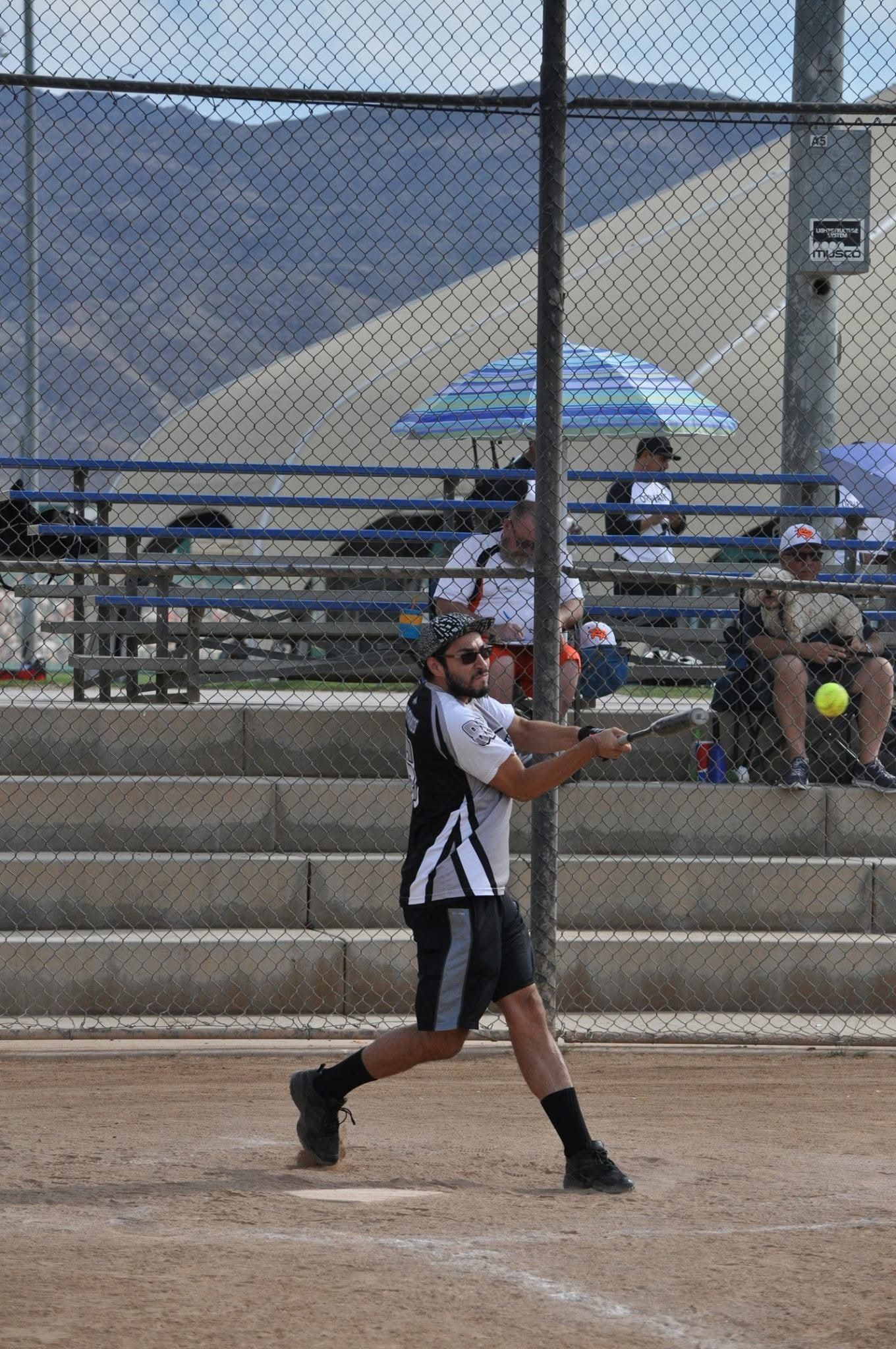 Softball player in black uniform swings a bat at home plate on a dirt field.