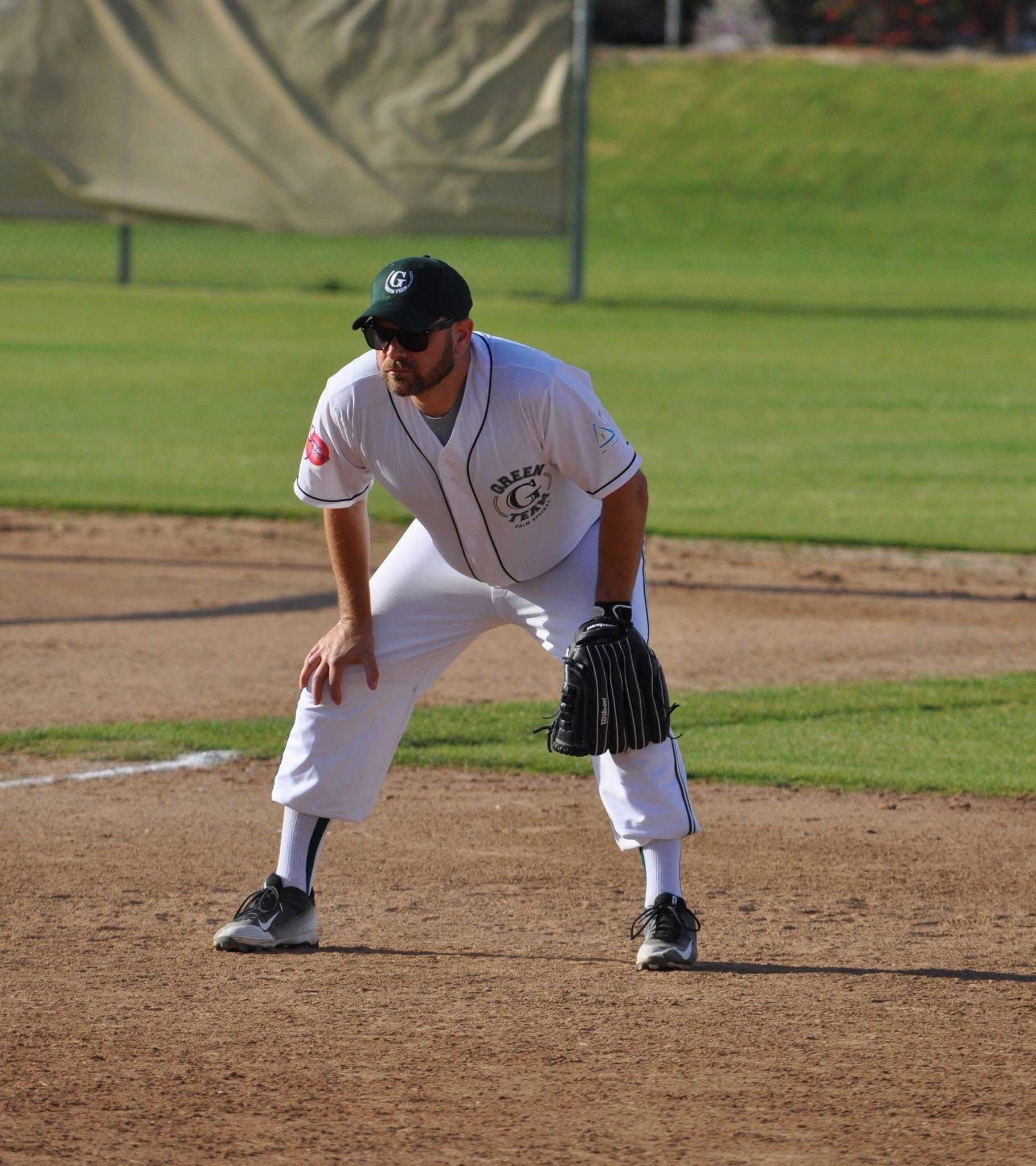 Baseball player in white uniform crouches on the infield, glove ready at a dirt diamond.