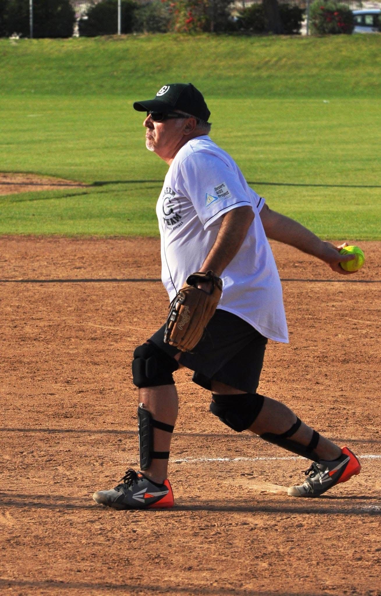 Softball pitcher in white shirt and black cap winds up on a dirt field with a yellow ball in hand.