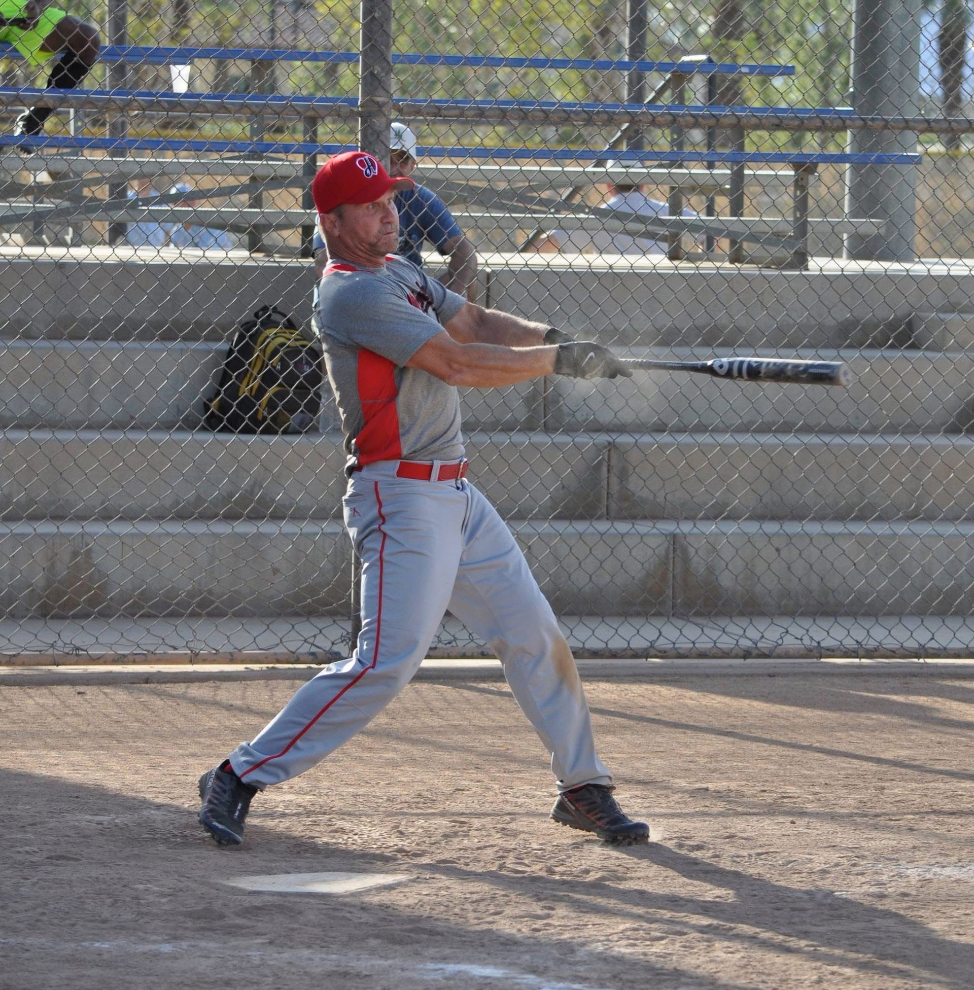 Baseball player in gray and red swinging a bat on a dirt field behind a chain-link fence