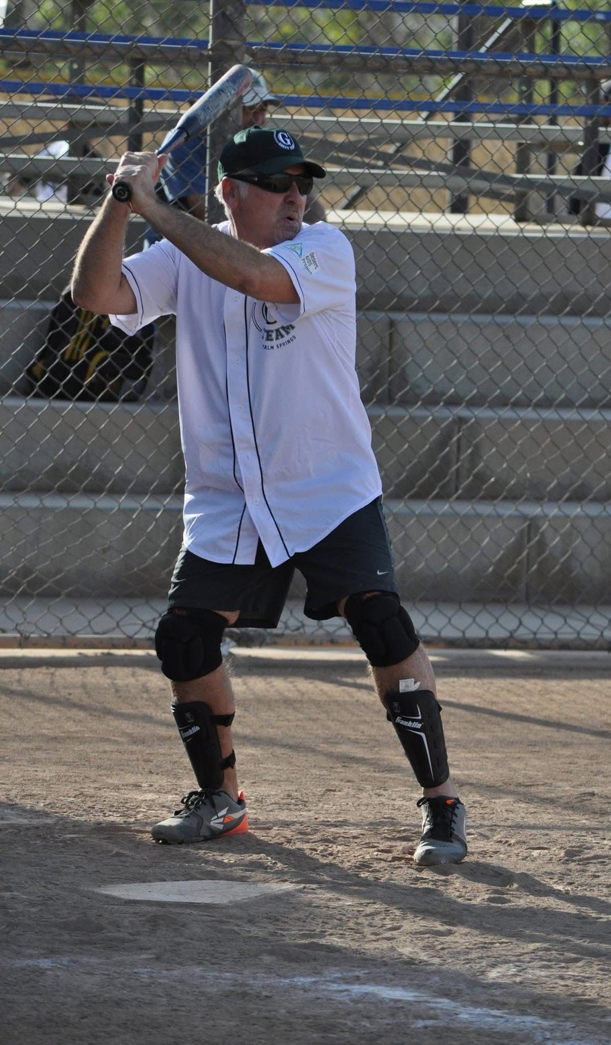Softball batter in white jersey and black helmet poised at the plate with bat raised, wearing knee braces.