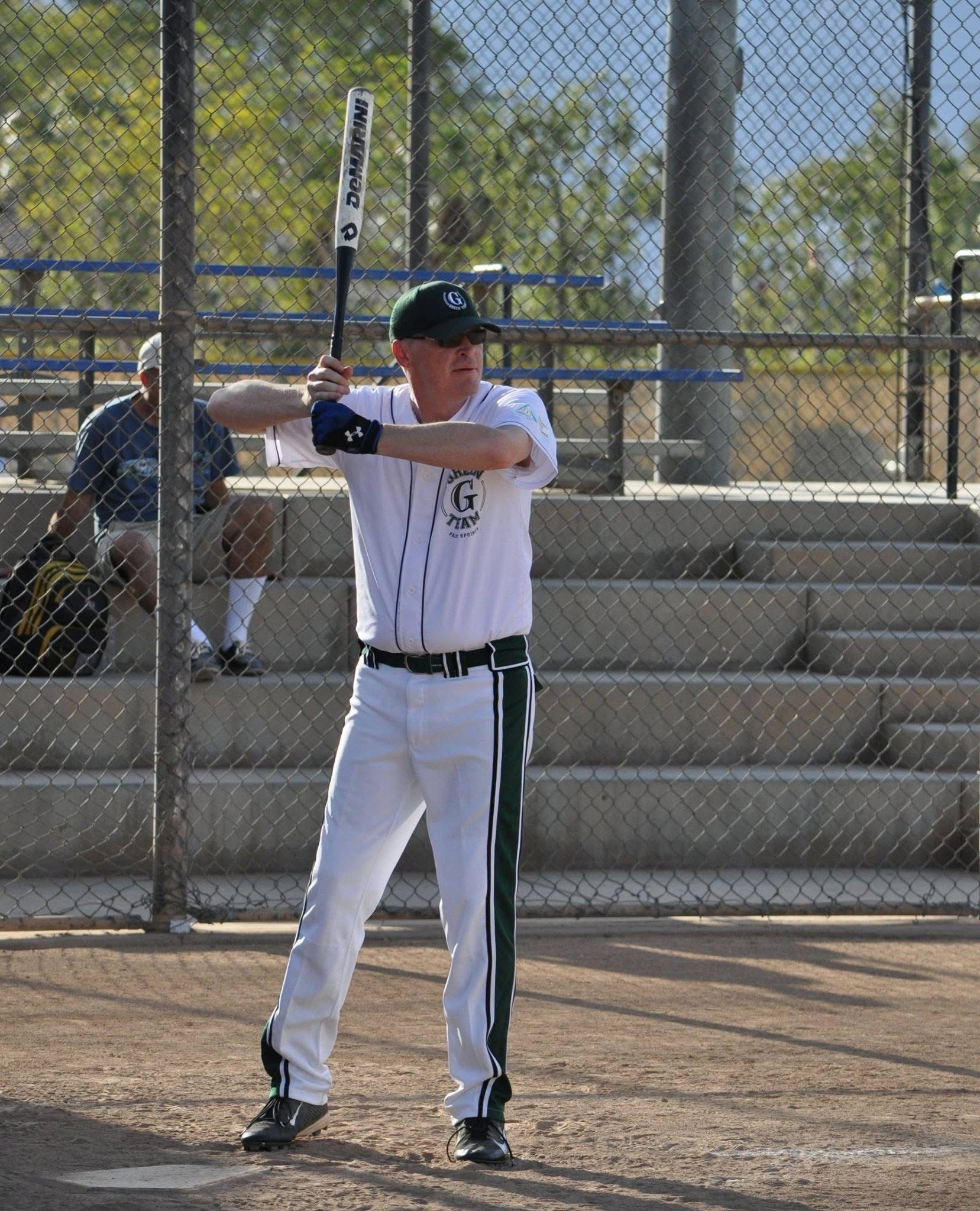 Baseball player in white uniform stands at bat on a field, holding a bat near a chain-link fence.