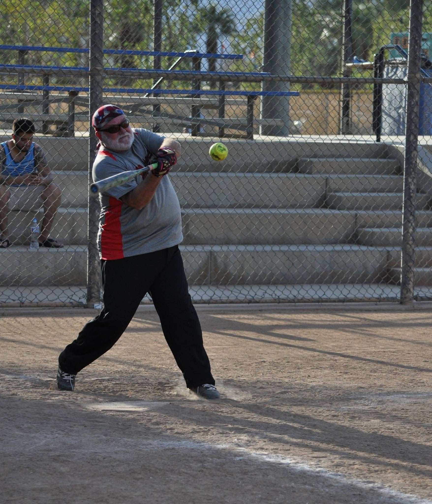 Softball player swings at a pitch on a dirt field, with bleachers and a chain-link fence behind.