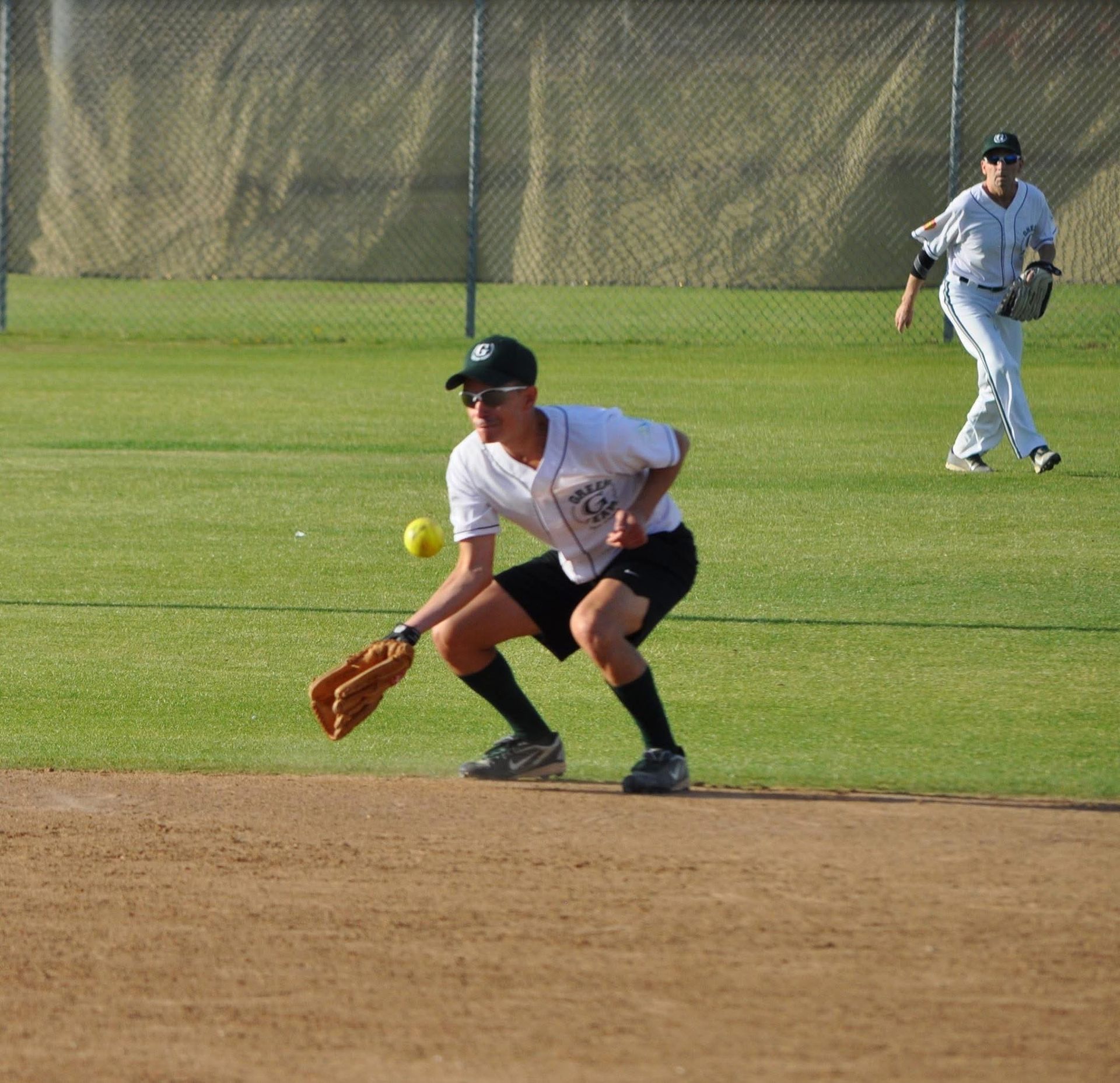 Baseball infielder in white uniform crouches on the infield as a ball rolls nearby, with an outfielder in the background