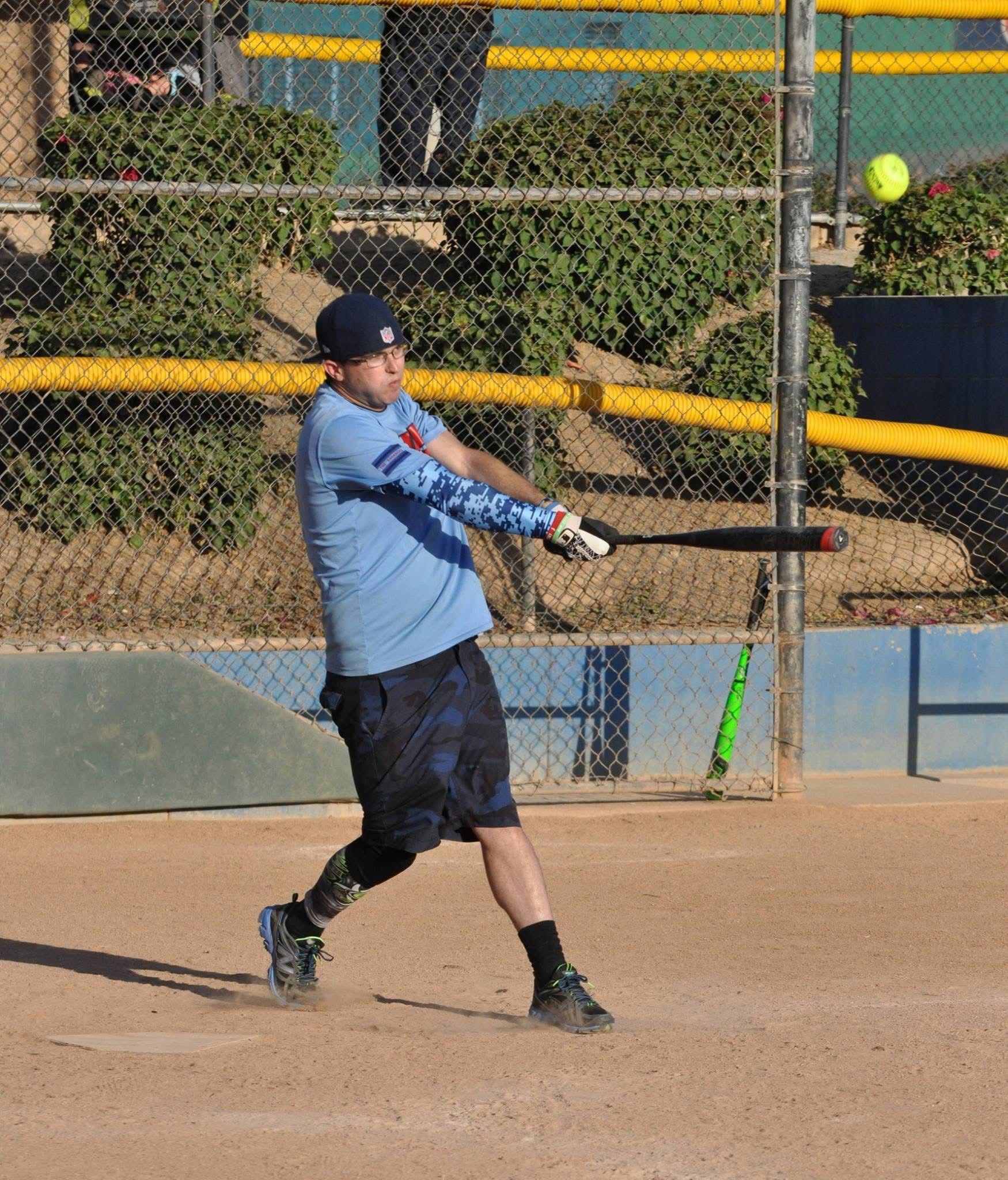 Baseball batter in blue swings at a pitch on a dirt field, with a chain-link fence and outfield behind him.