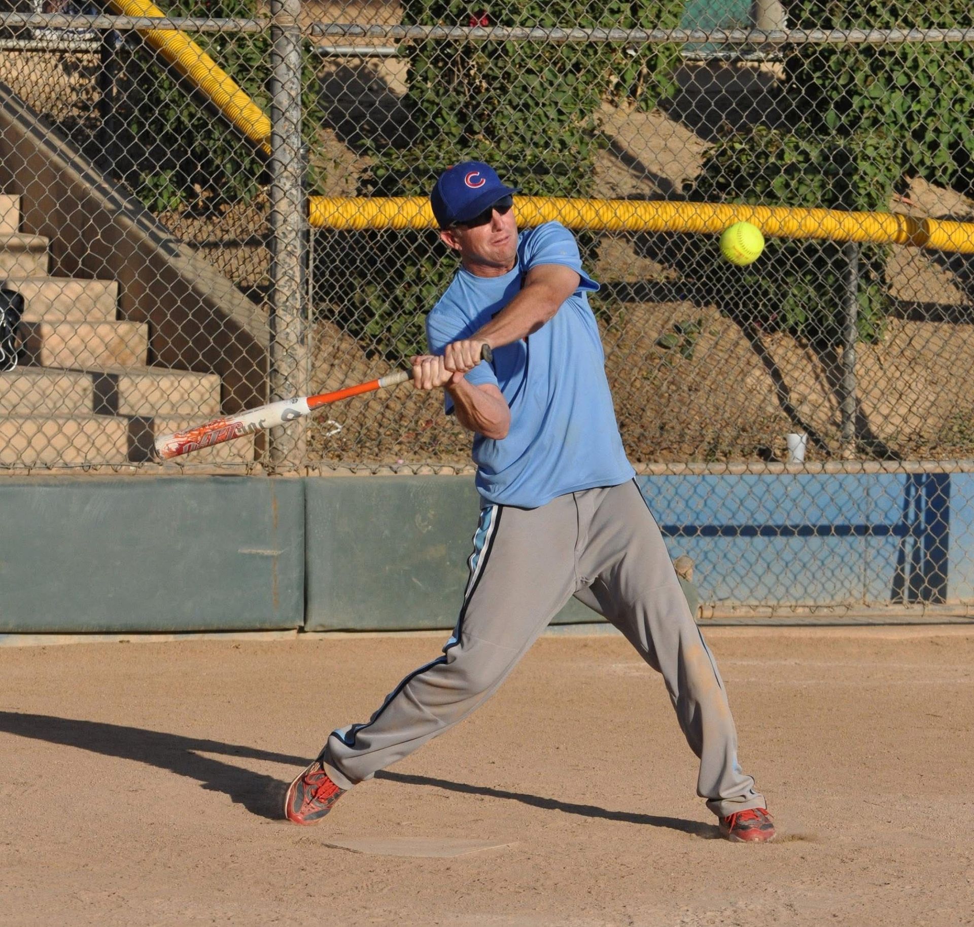 Baseball player in blue swings a bat on a dirt field near a chain-link fence.