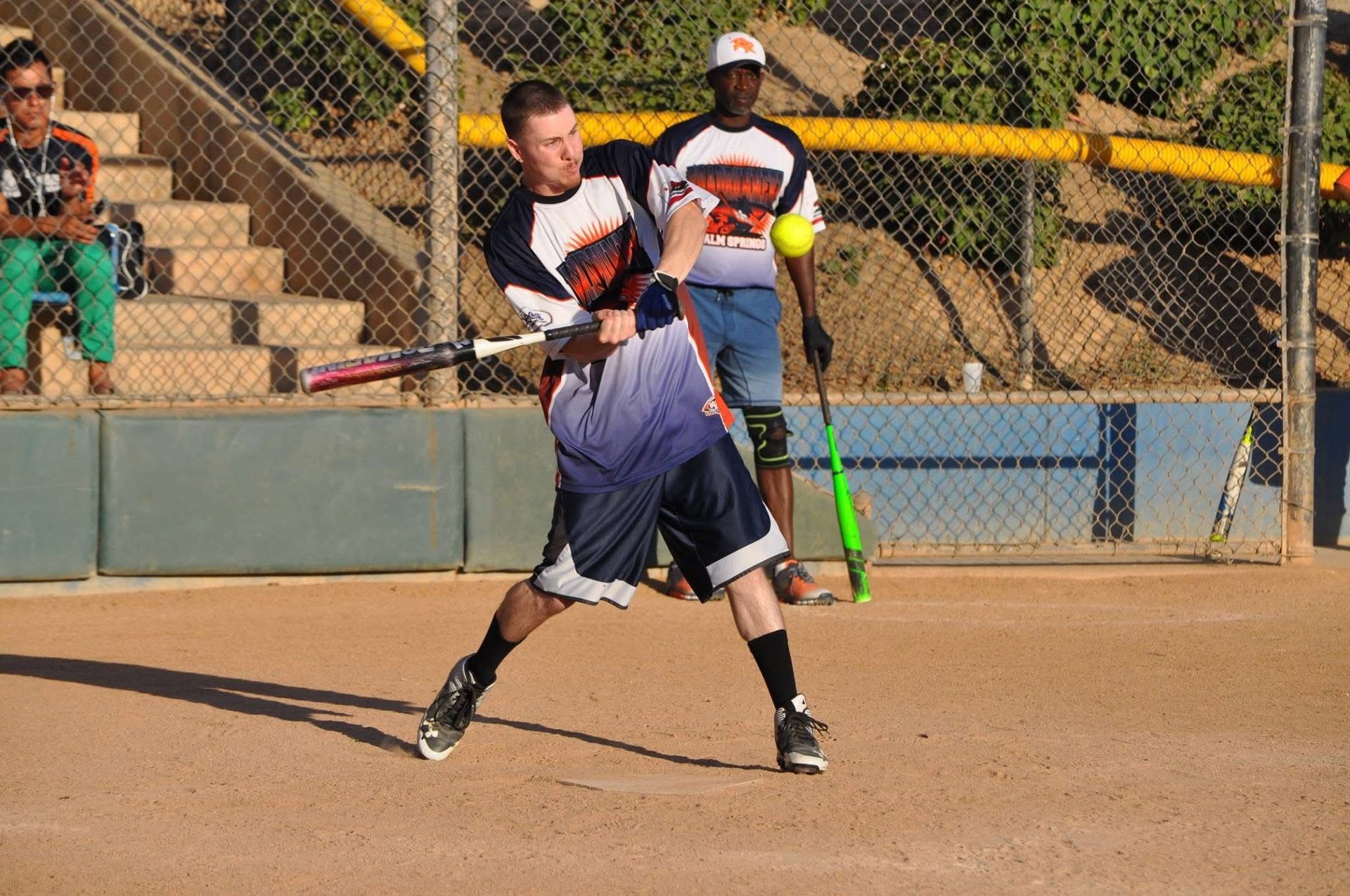 Softball batter swinging at a yellow ball on a dirt field with a fence and spectators behind him