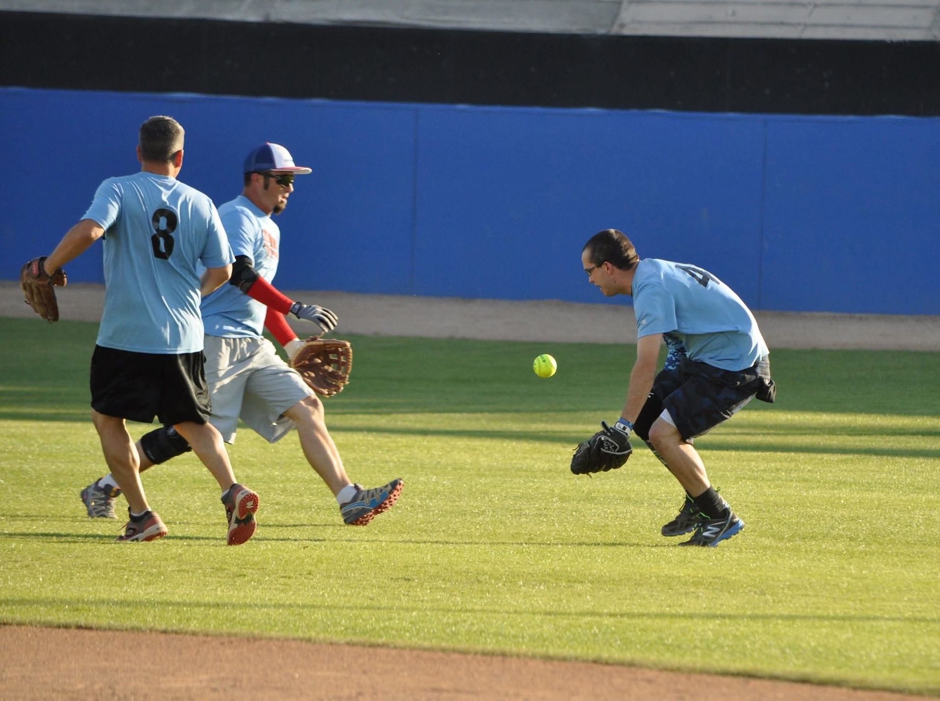 Three baseball players in light blue uniforms field a ground ball on a sunlit grass field.