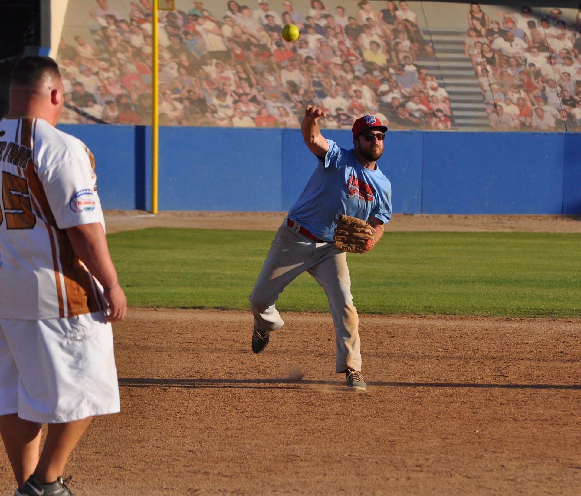 Baseball pitcher in blue uniform throwing a pitch on a dirt field, with a batter watching nearby.