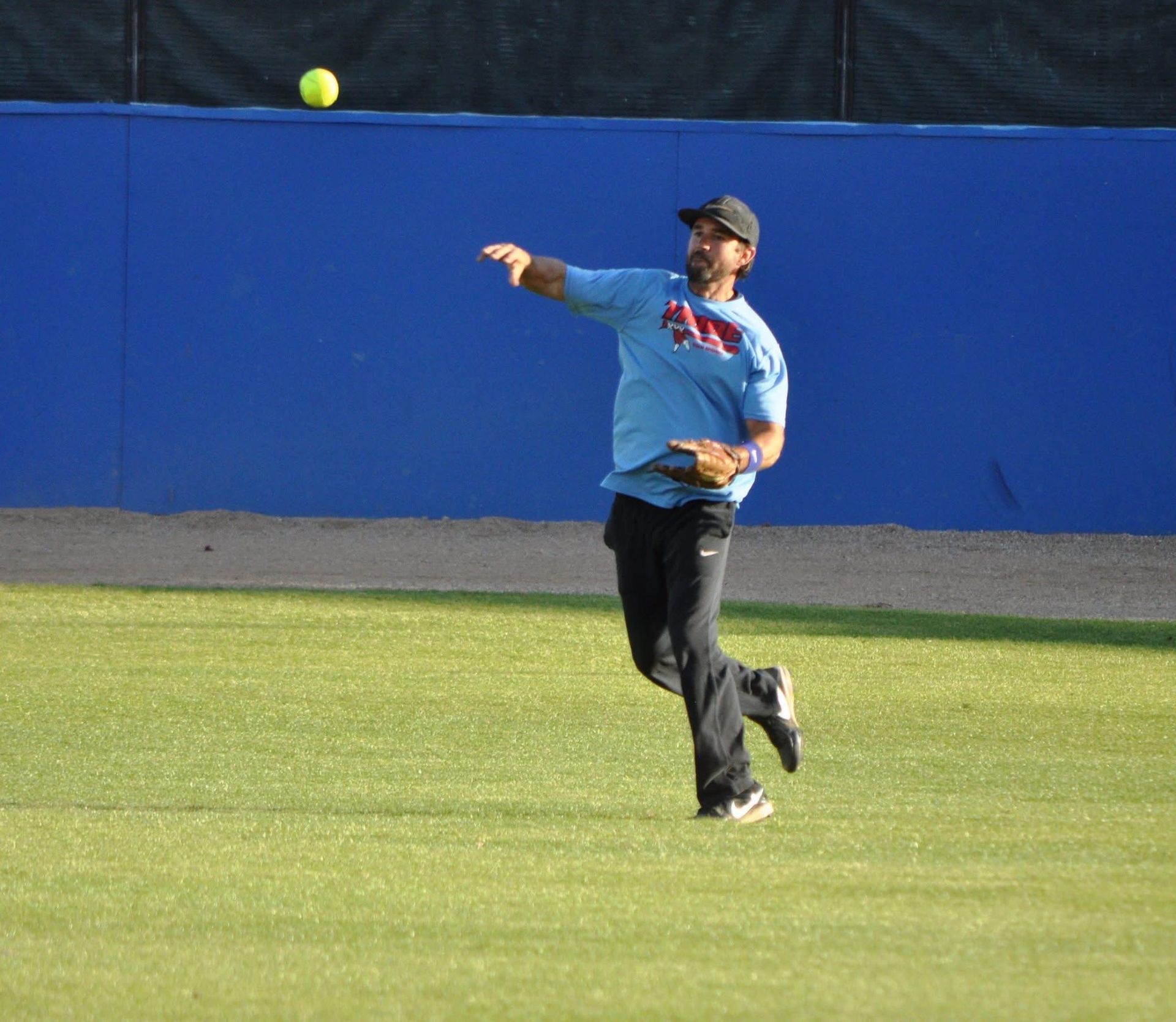 Baseball player in blue shirt reaching for a ball on a grassy field near the outfield wall.