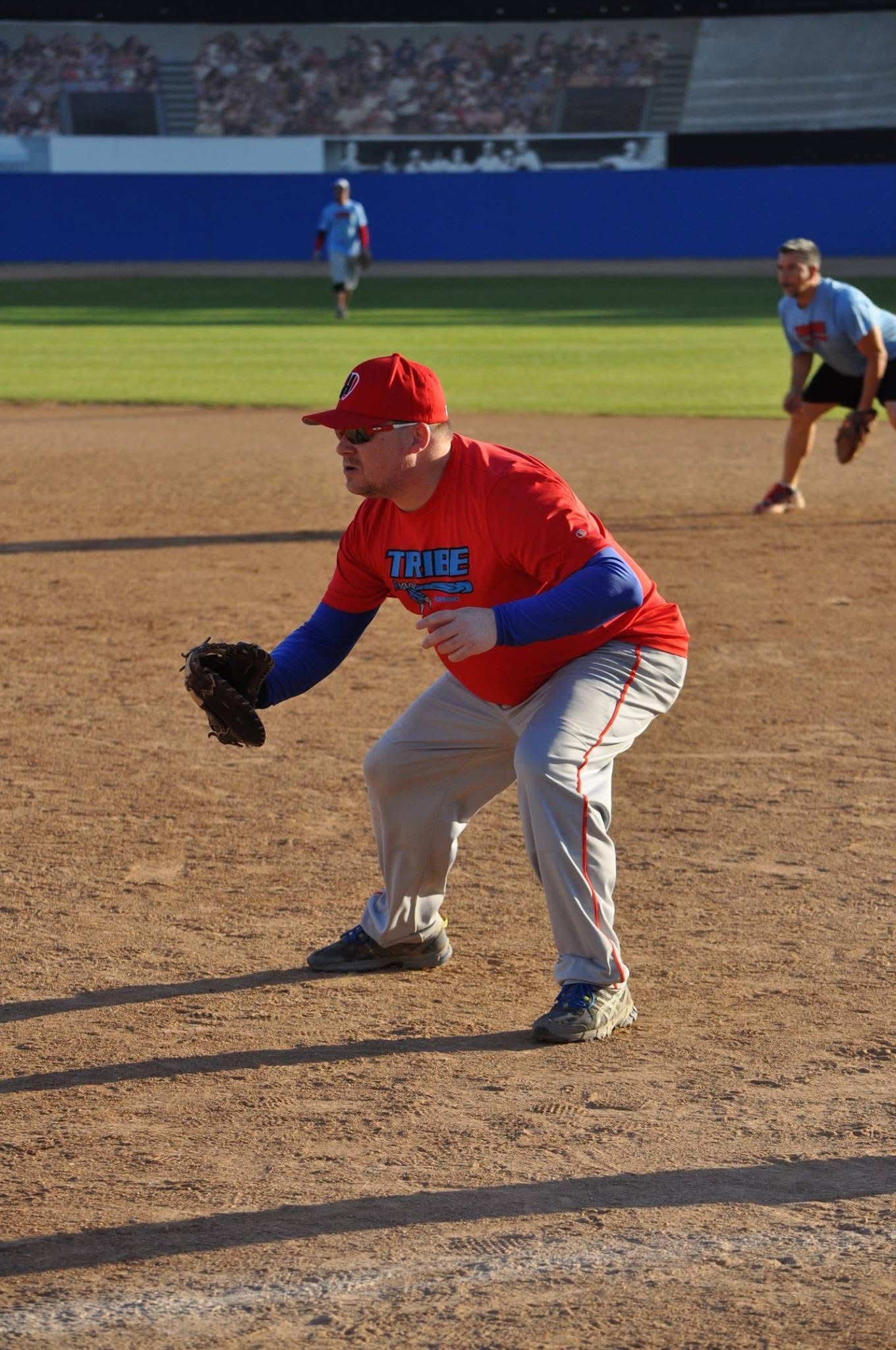 Baseball infielder in red and blue crouches to field a ground ball on the dirt infield.