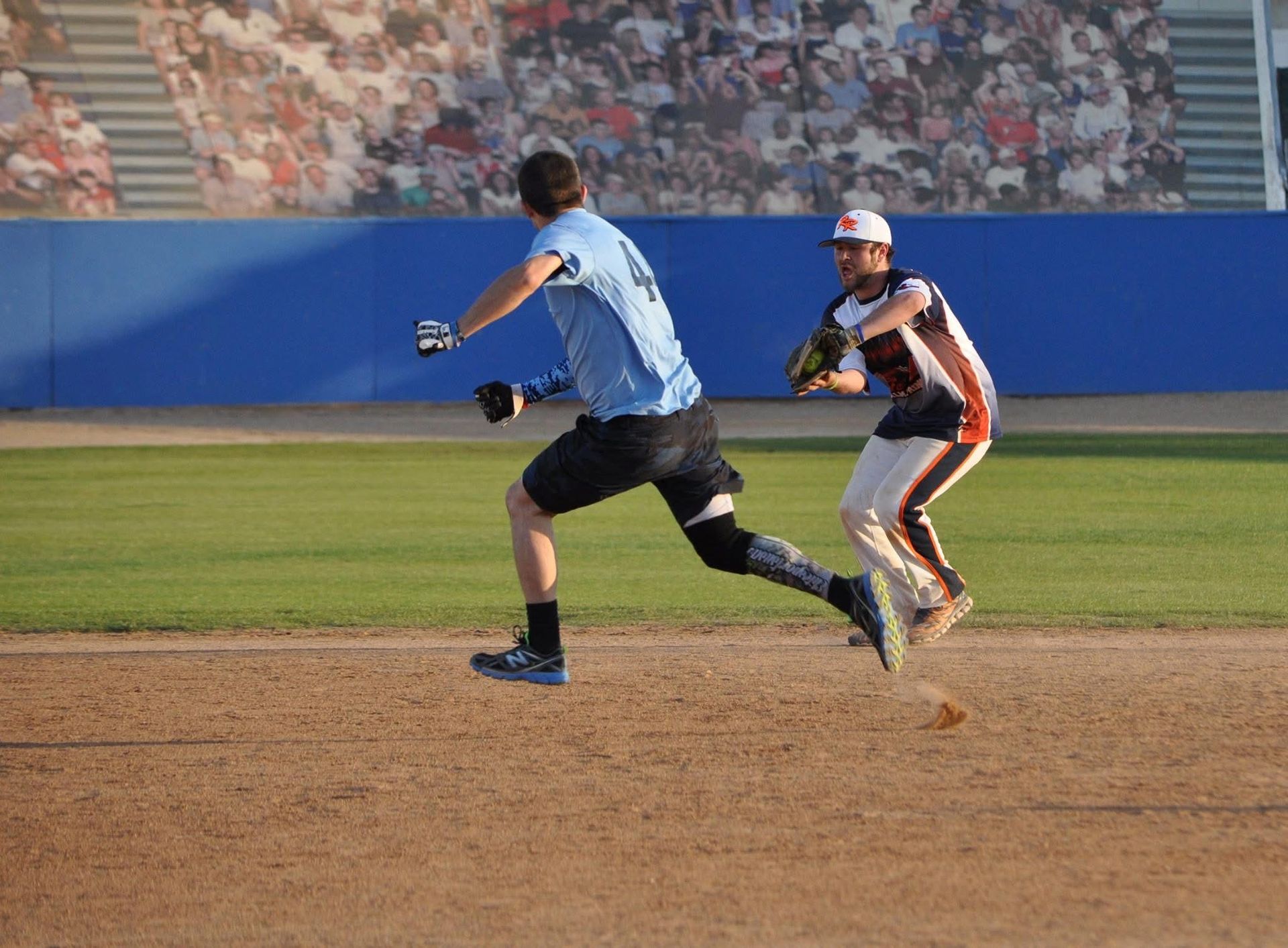 Two baseball players sprint on a dirt infield, one in blue sliding toward a catch by a fielder in white and black.