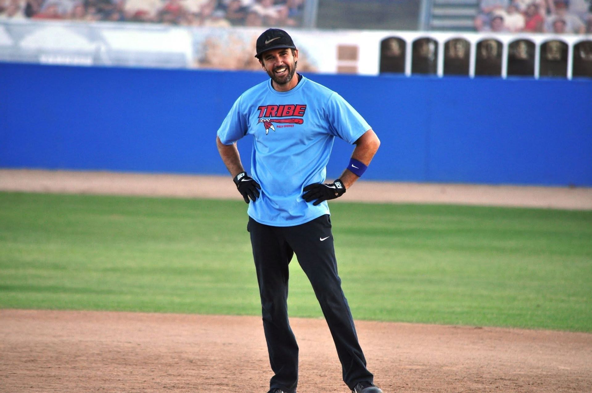 Man standing on a baseball field in a light blue shirt and black pants, hands on hips.
