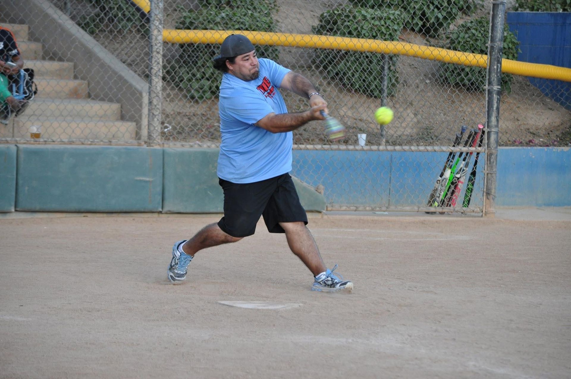 Softball player in blue shirt swings at a yellow ball on a dirt field