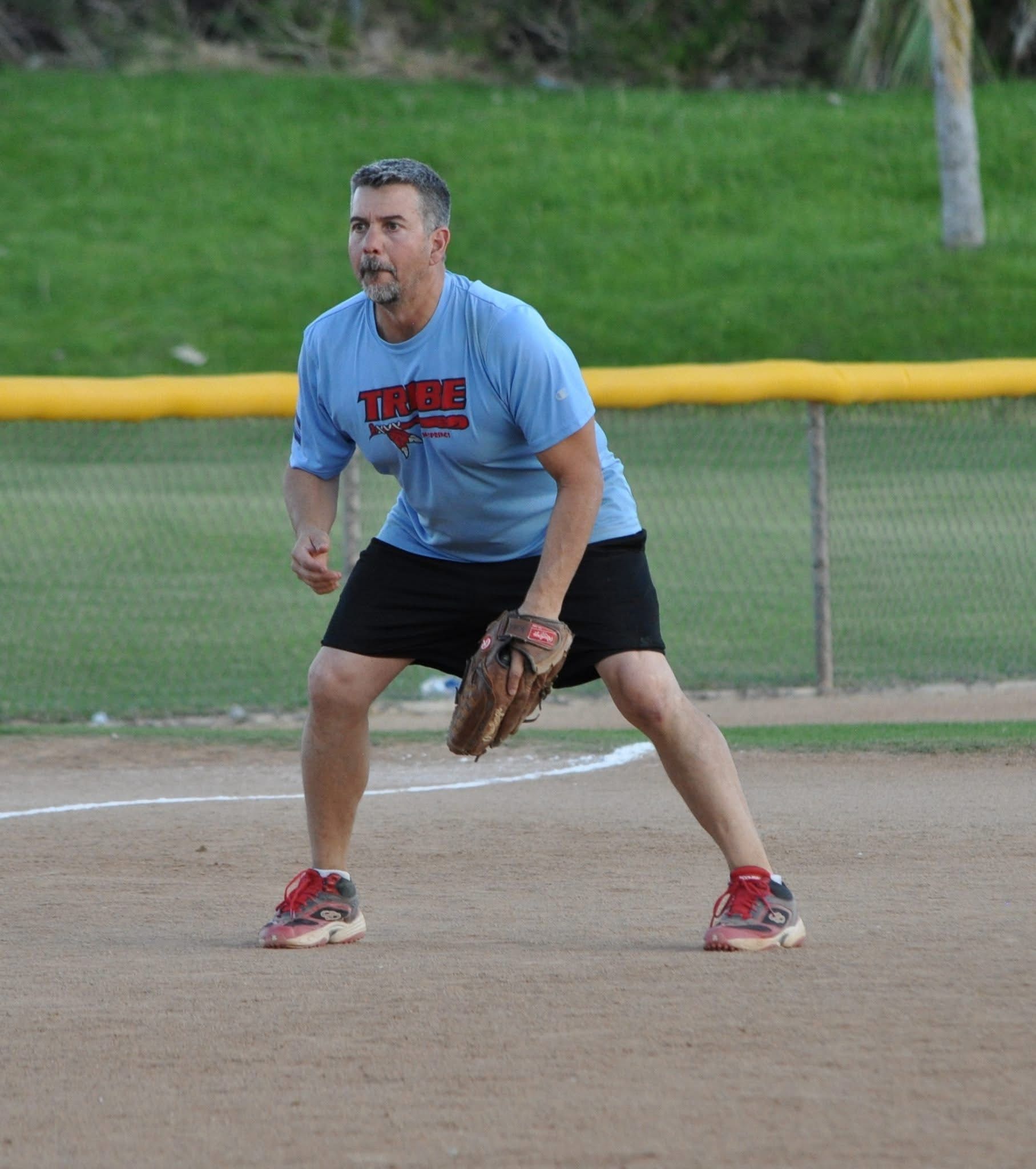Man in blue shirt and black shorts crouches on a baseball field, ready to field a ball.