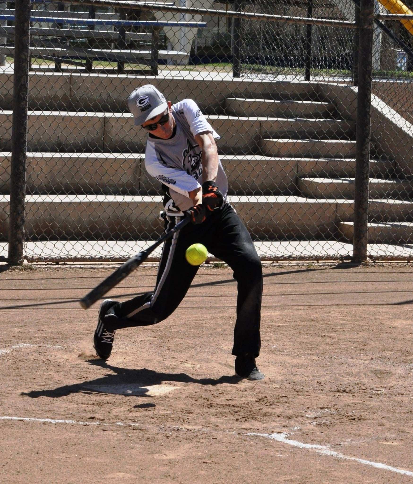 Softball player in white swings at a yellow ball on a dirt field near bleachers and chain-link fence
