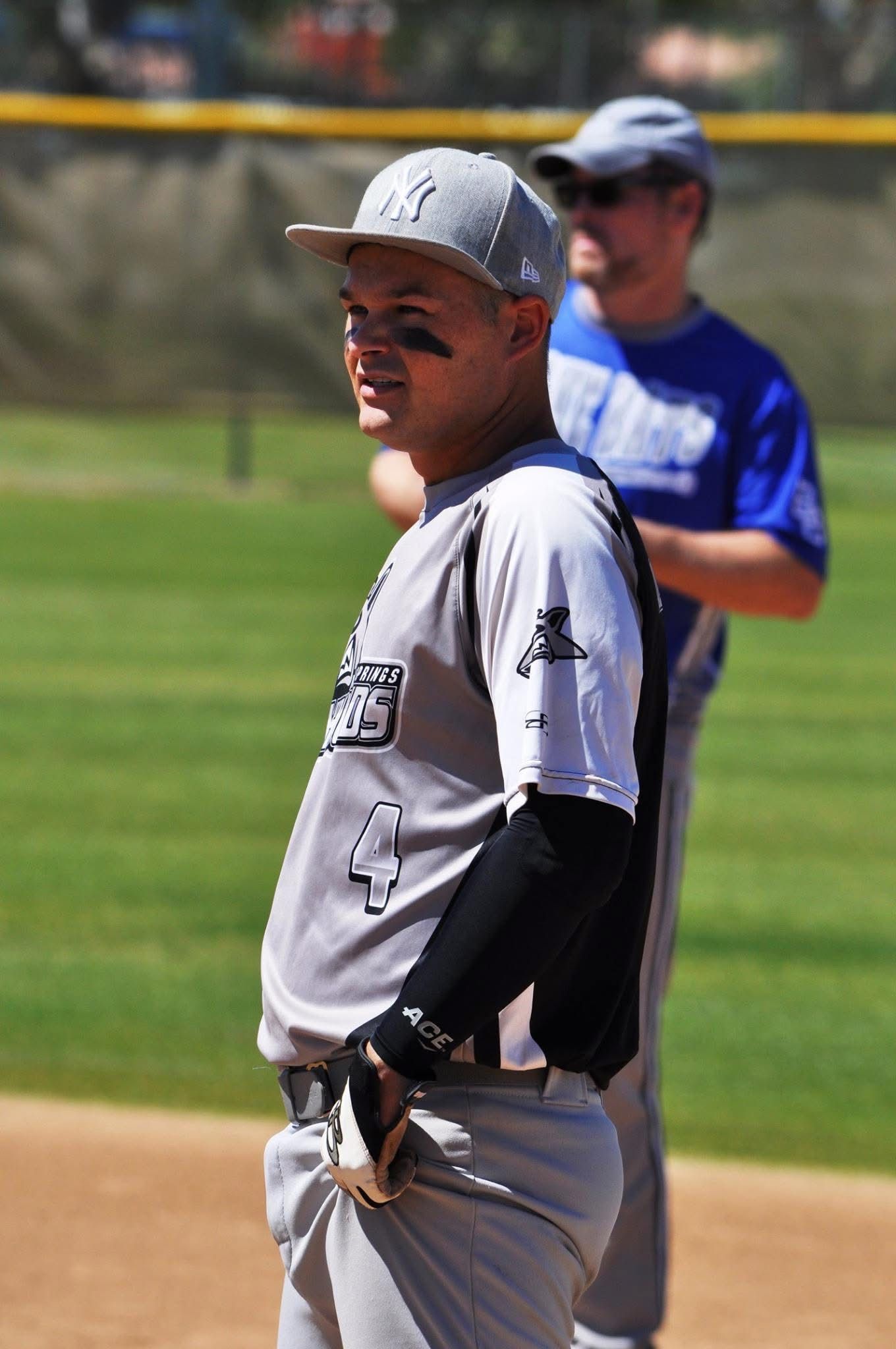 Baseball player in gray uniform and cap standing on the field, with a blurred player in blue behind him.