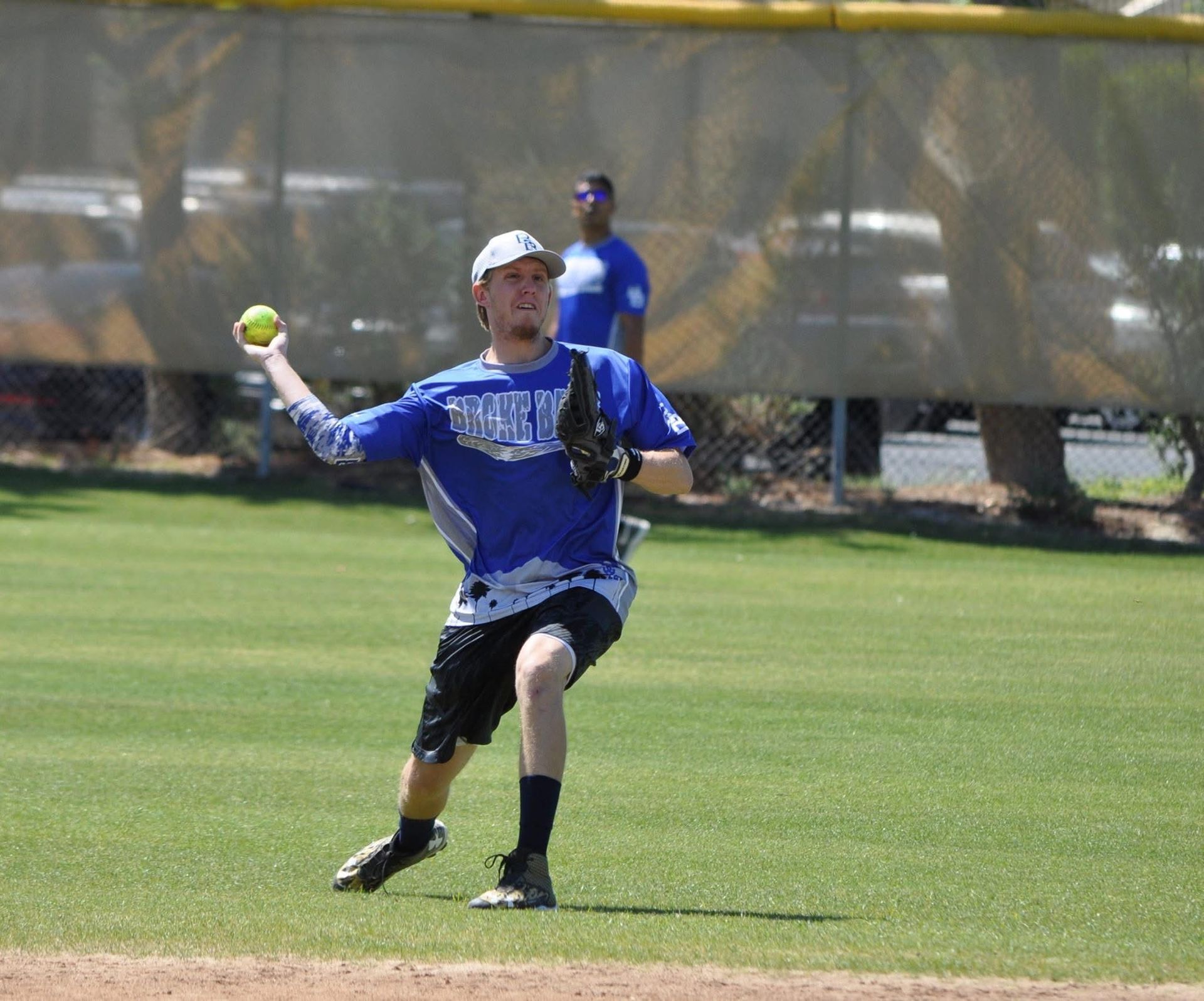 Softball player in blue jersey and white cap throwing a ball on a grassy field
