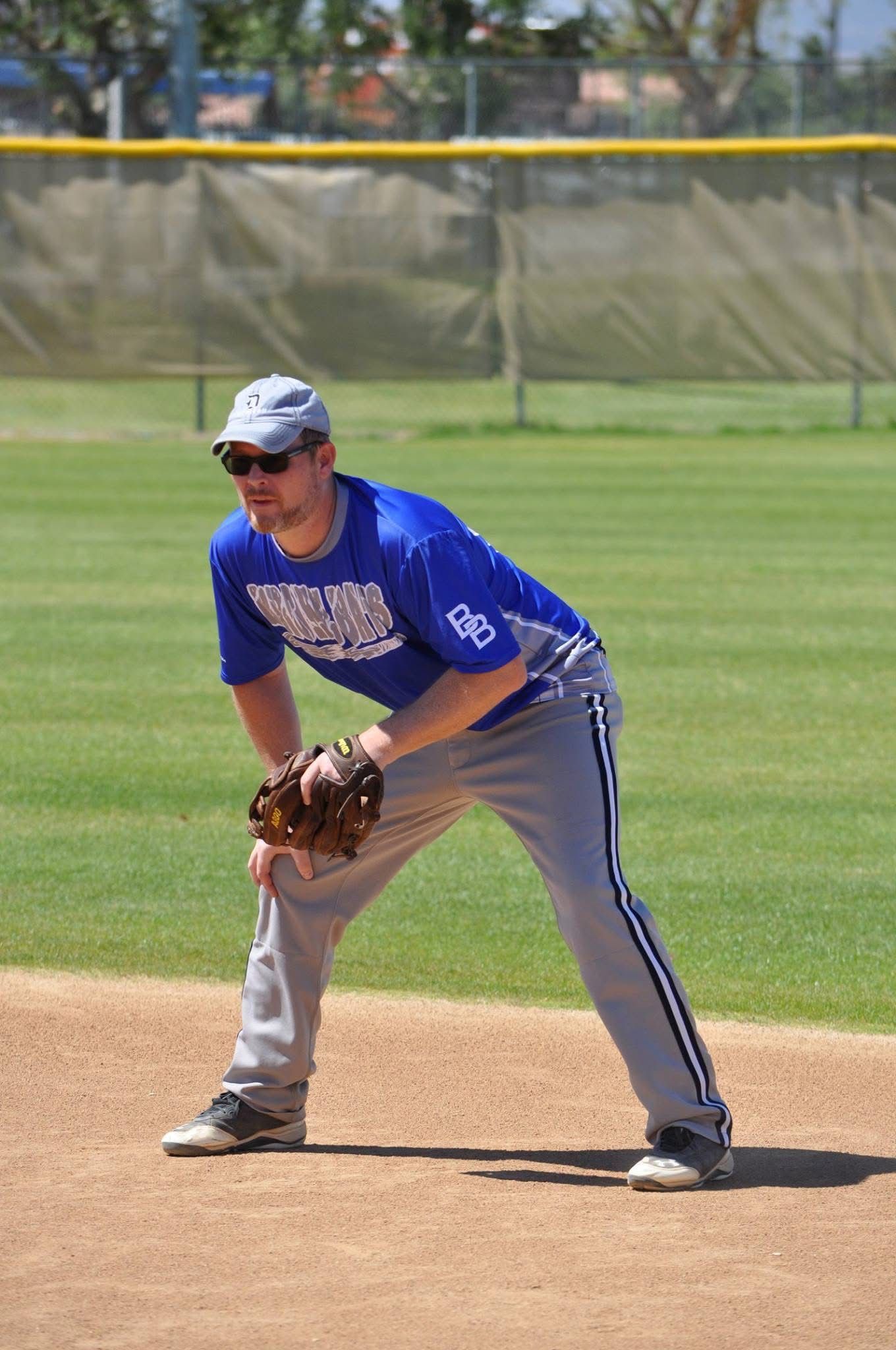 Baseball infielder in blue uniform crouching on the infield dirt, glove ready, with outfield fence behind.