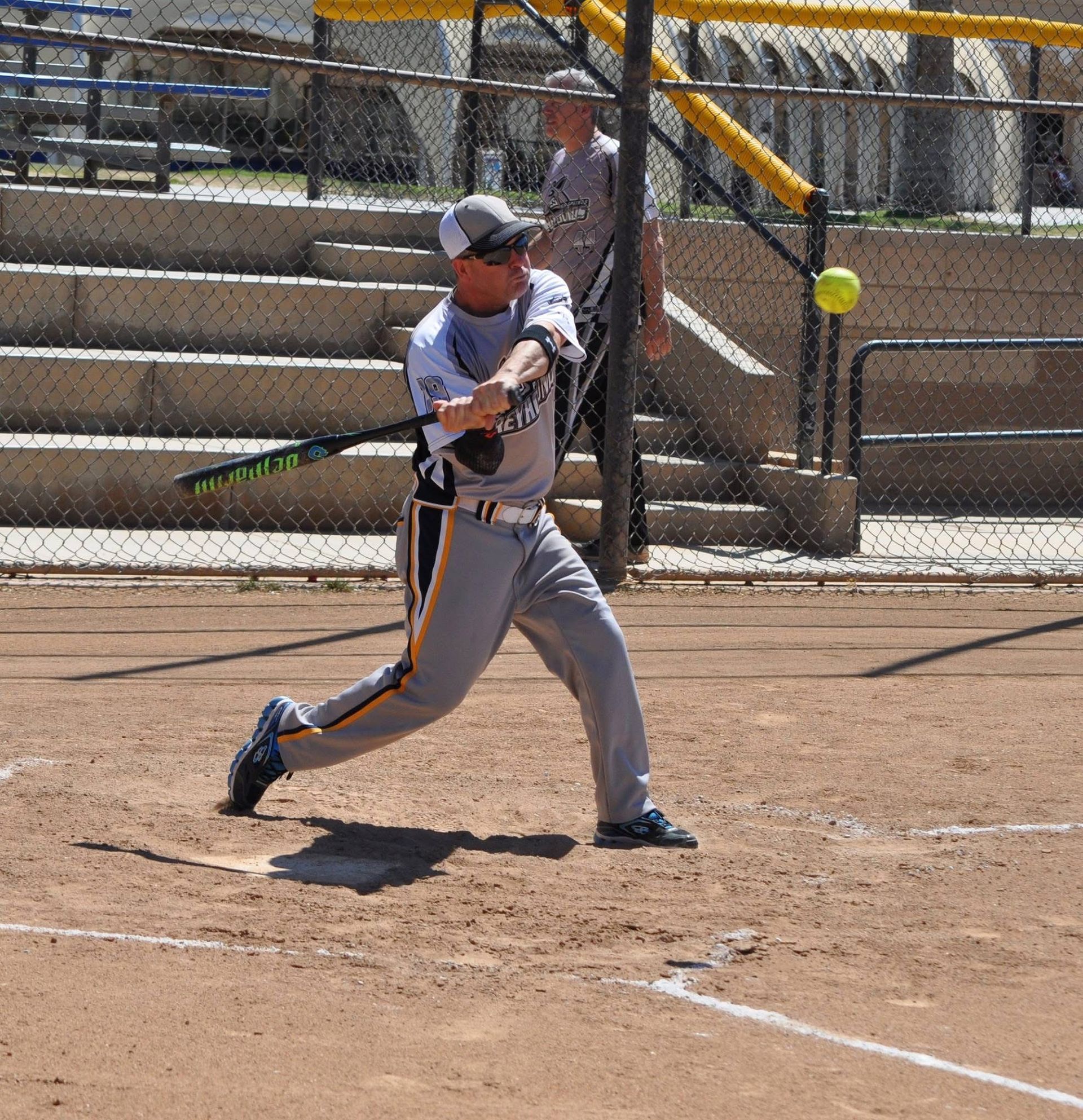 Softball batter in gray uniform swings at a pitch on a dirt field behind a chain-link fence.