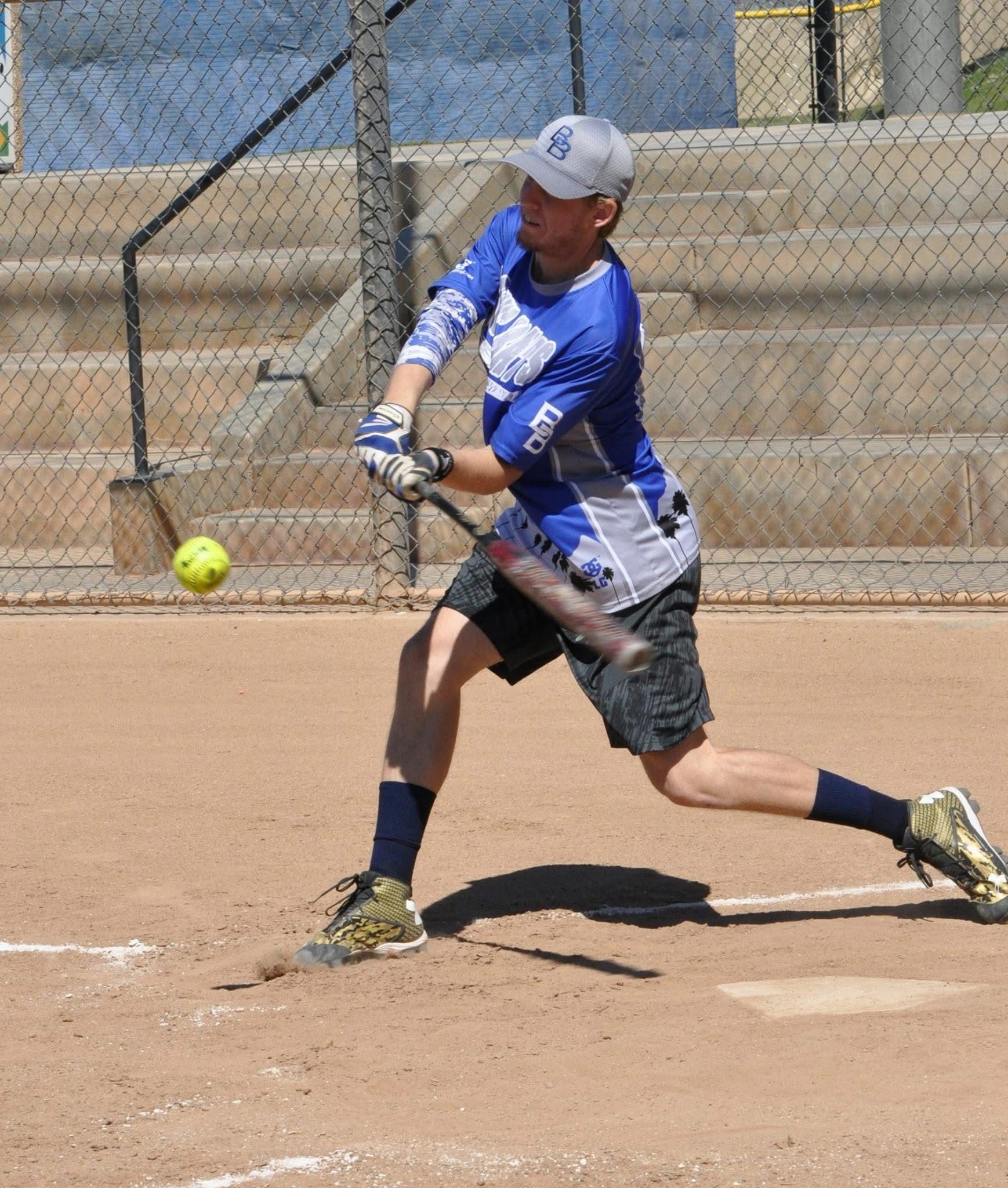 Softball player in blue swings at a pitch on a dirt field during a game