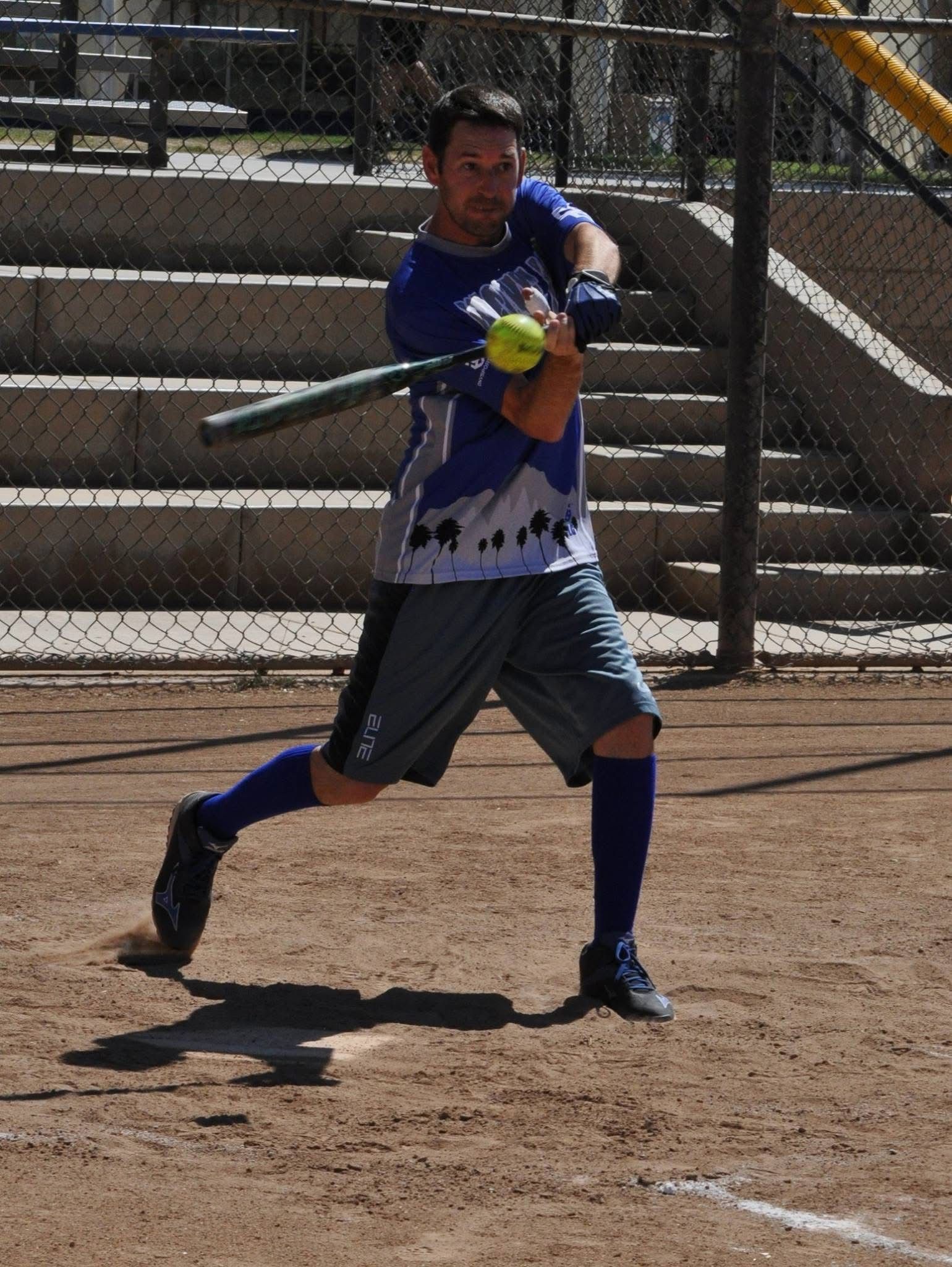 Softball batter in blue swings at a pitch on a dirt field near concrete bleachers.