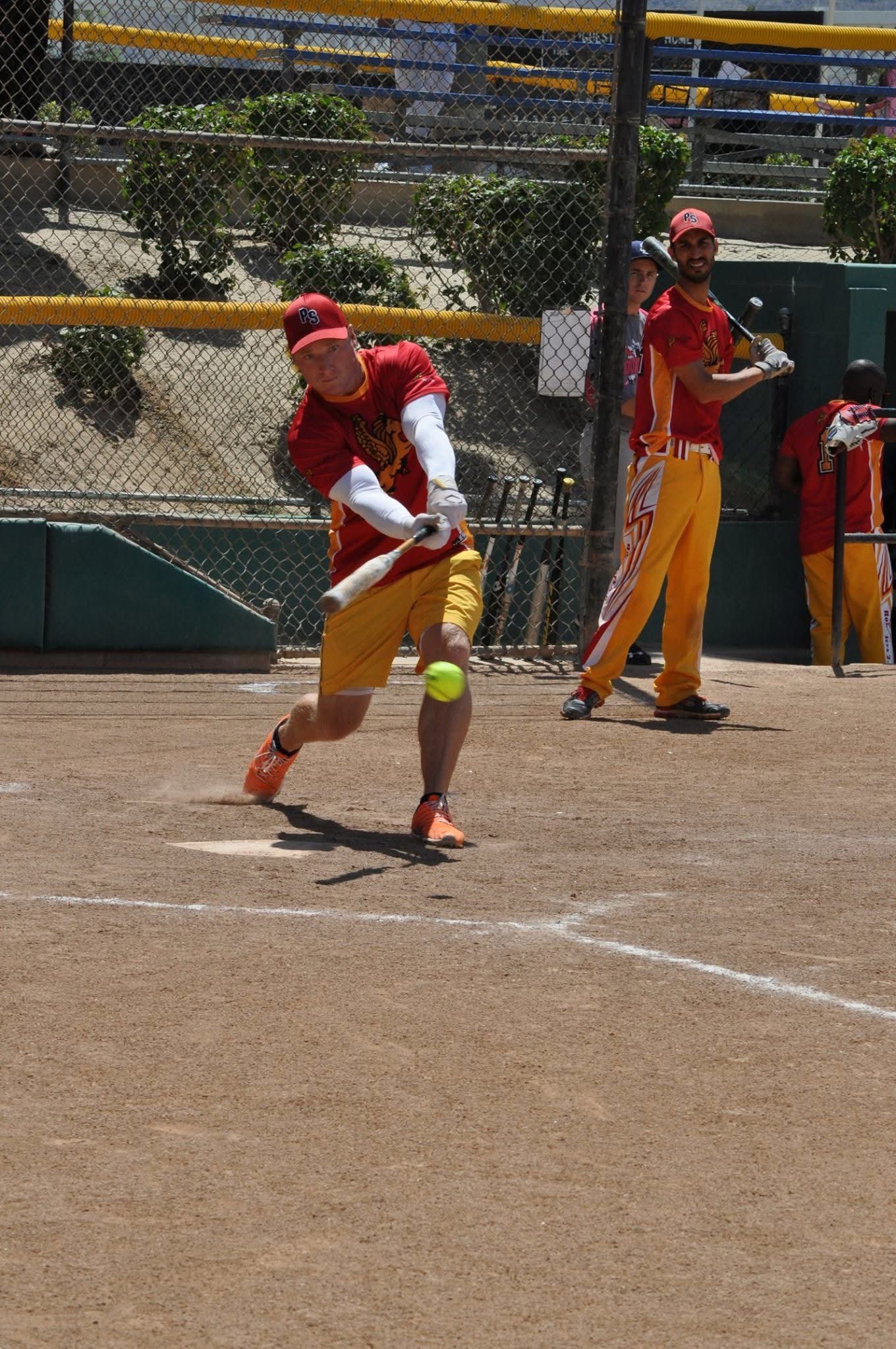 Softball batter in red uniform swings at a pitch on a dirt field with teammates and fence behind him.
