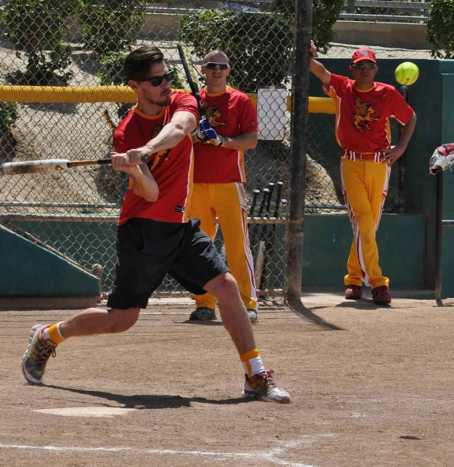 Softball batter in red swings at a pitch while teammates watch near the fence.