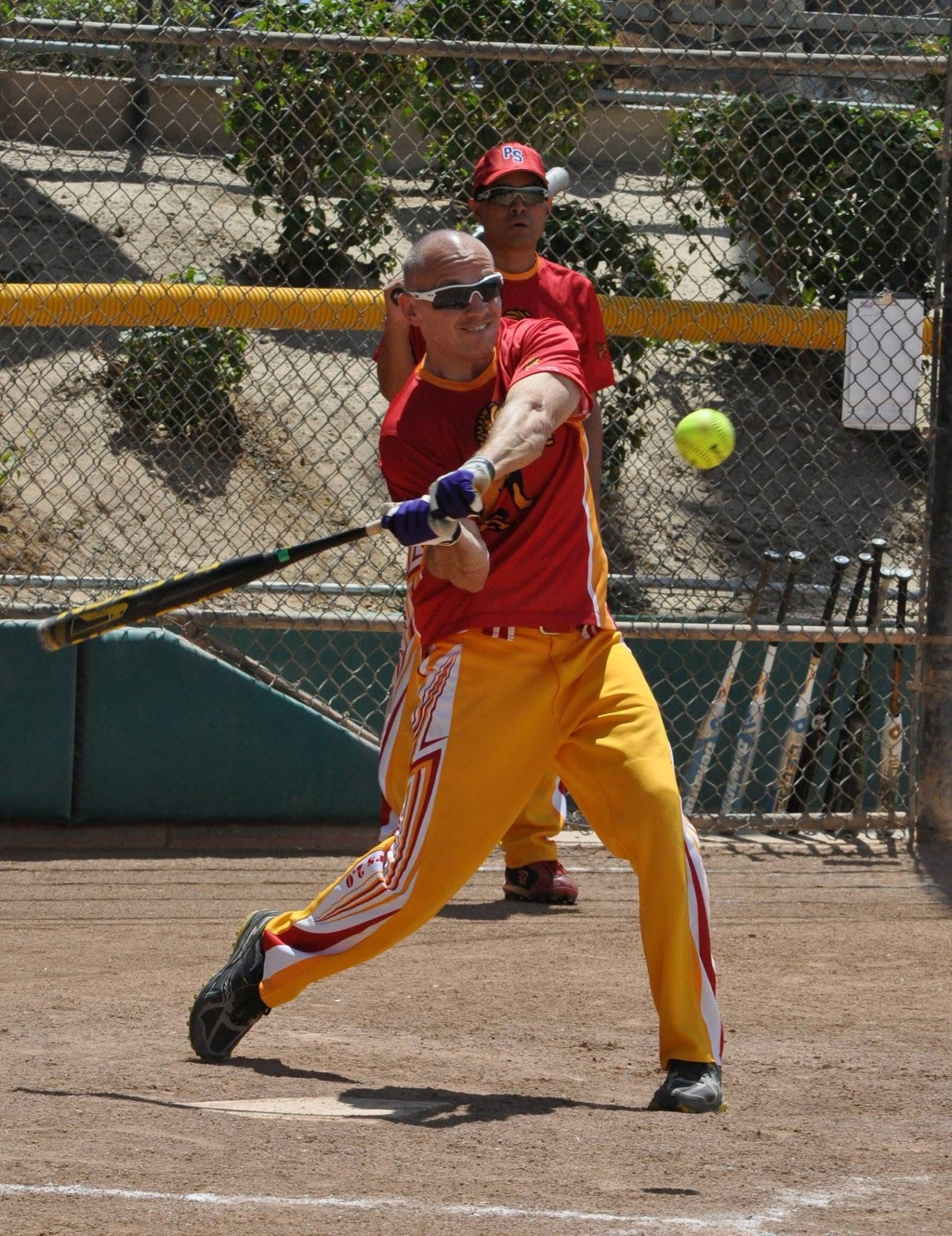 Softball batter in red and yellow swings at a pitch on a dirt field, with catcher behind him.