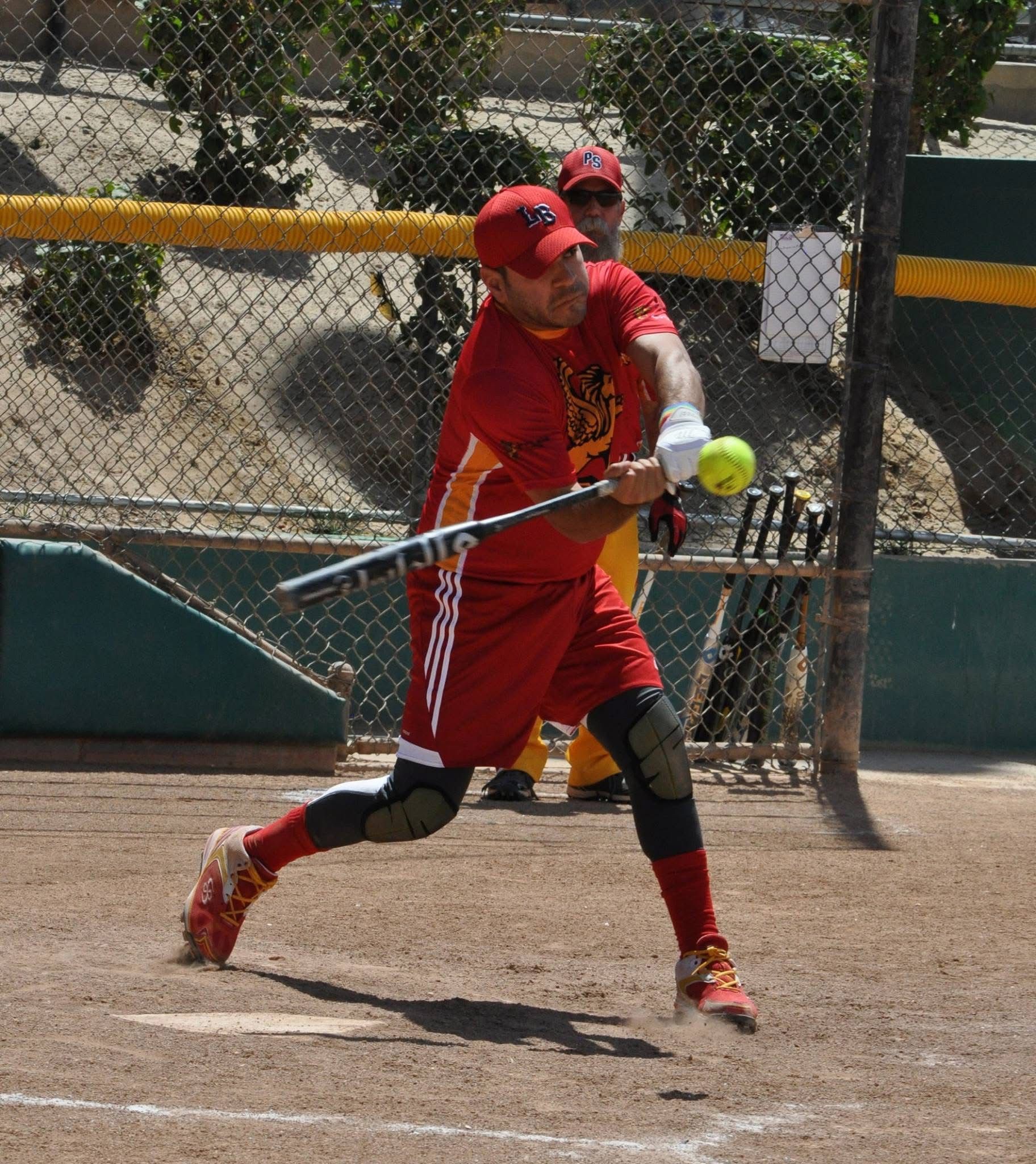 Softball player in red uniform swinging at a pitch on a dirt field, bat and ball visible.