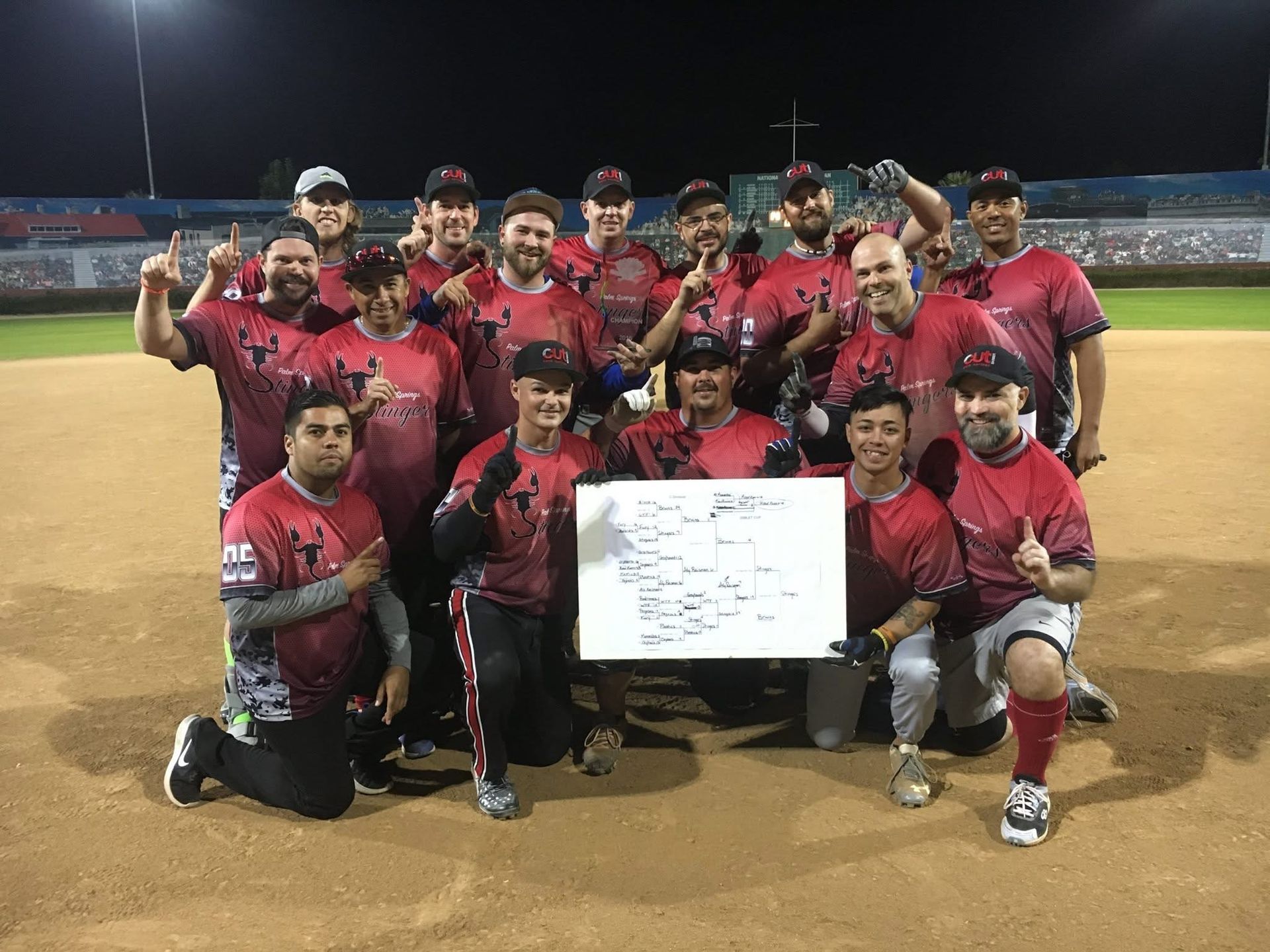 Baseball team posing on the field at night with a white sign, wearing red jerseys and black caps