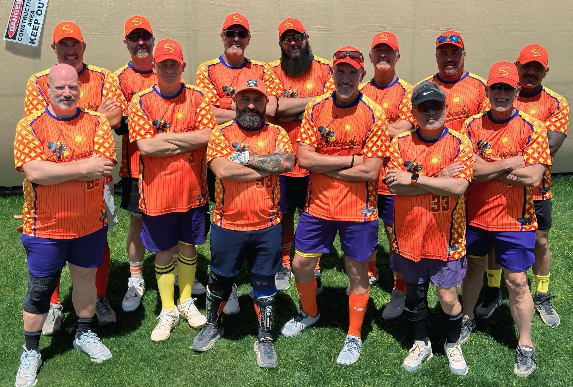 Cricket team in orange uniforms posing together on a grassy field, arms crossed and smiling
