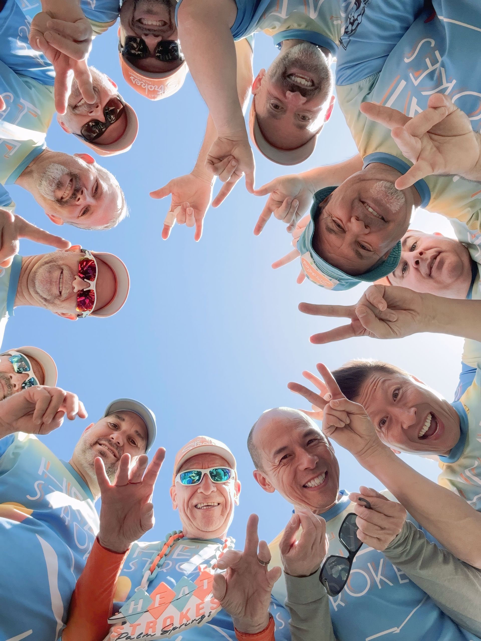 Group of smiling people leaning in a circle outdoors, hands raised toward the camera against a blue sky.