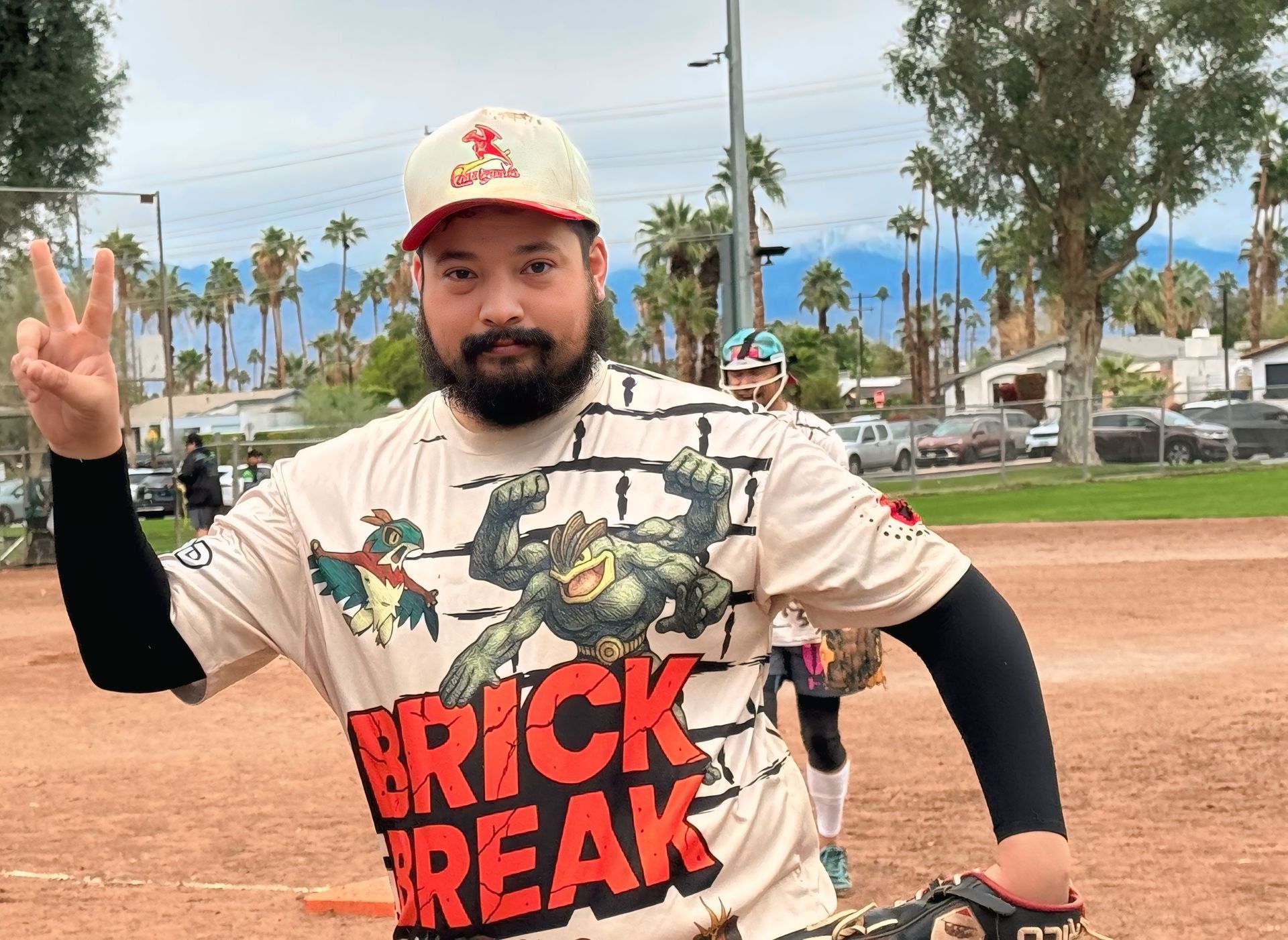 Man in a graphic T-shirt and cap flashes a peace sign on a dirt path at an outdoor park.
