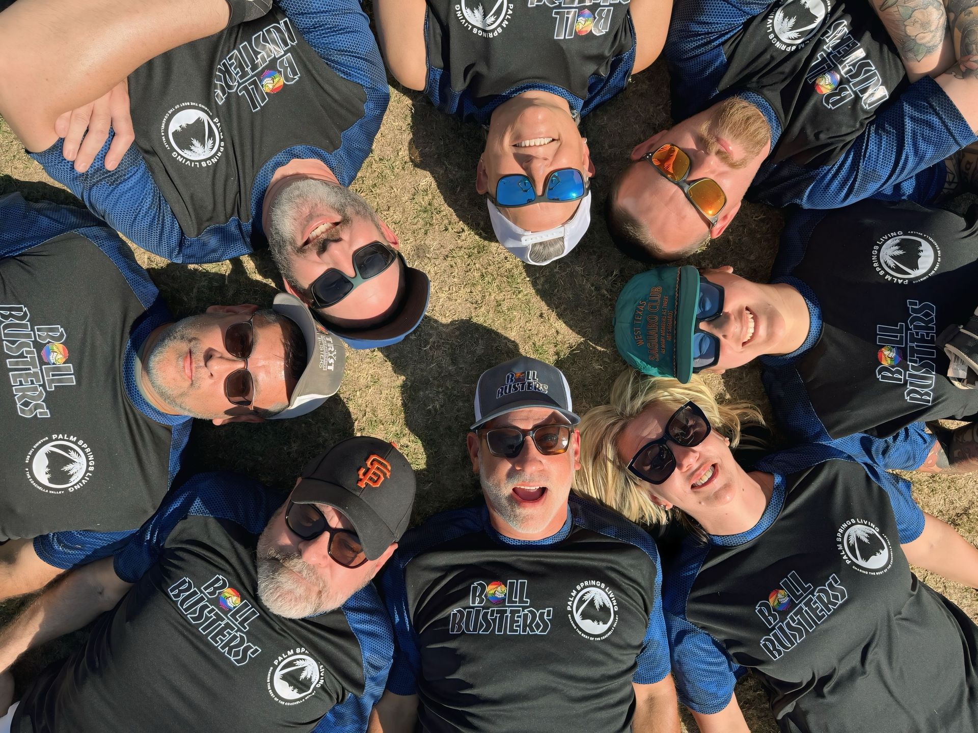 People in black shirts and sunglasses lying in a circle on grass, smiling up at the camera