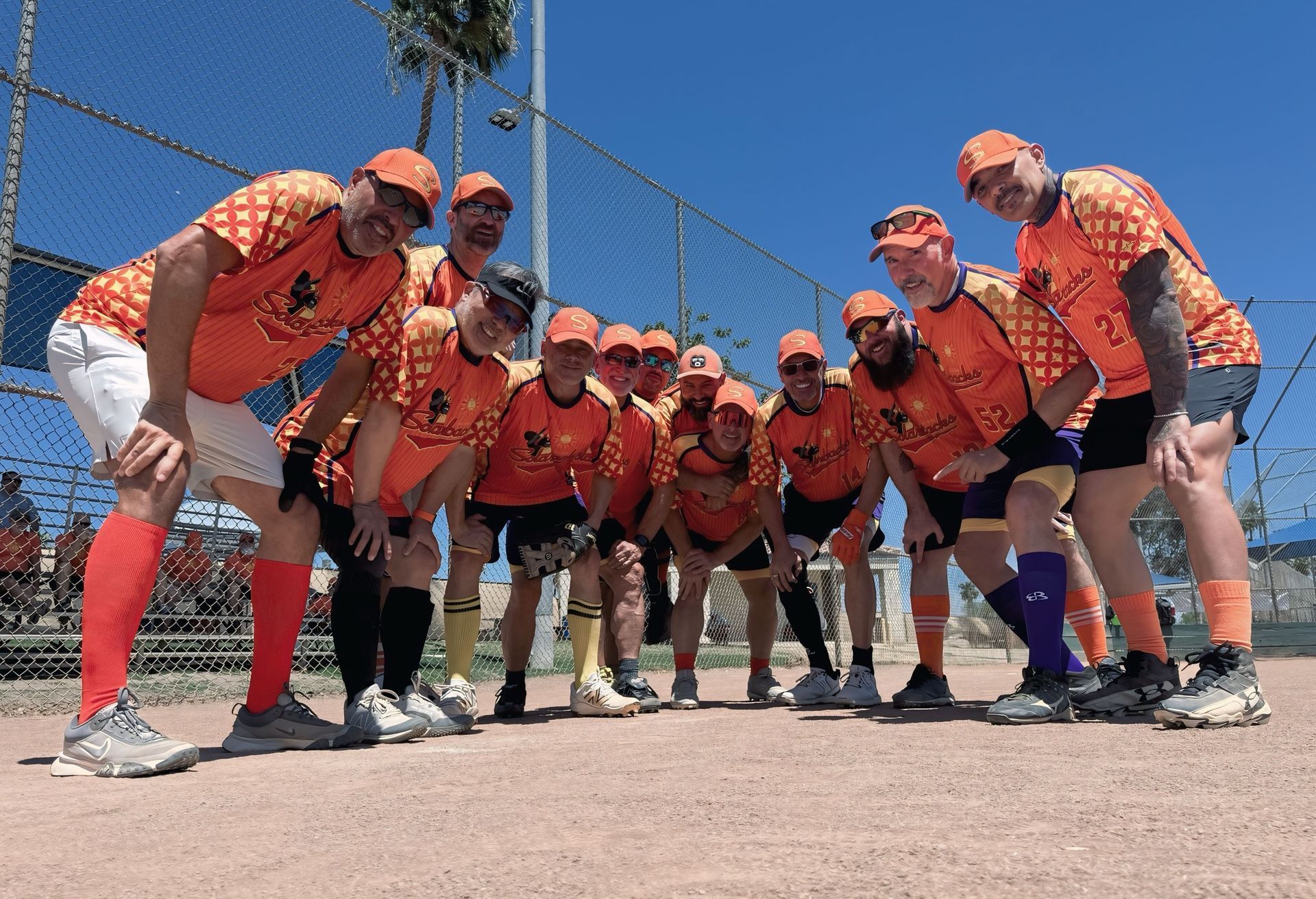 Group of softball players in orange jerseys posing in a low crouch on a sunny field