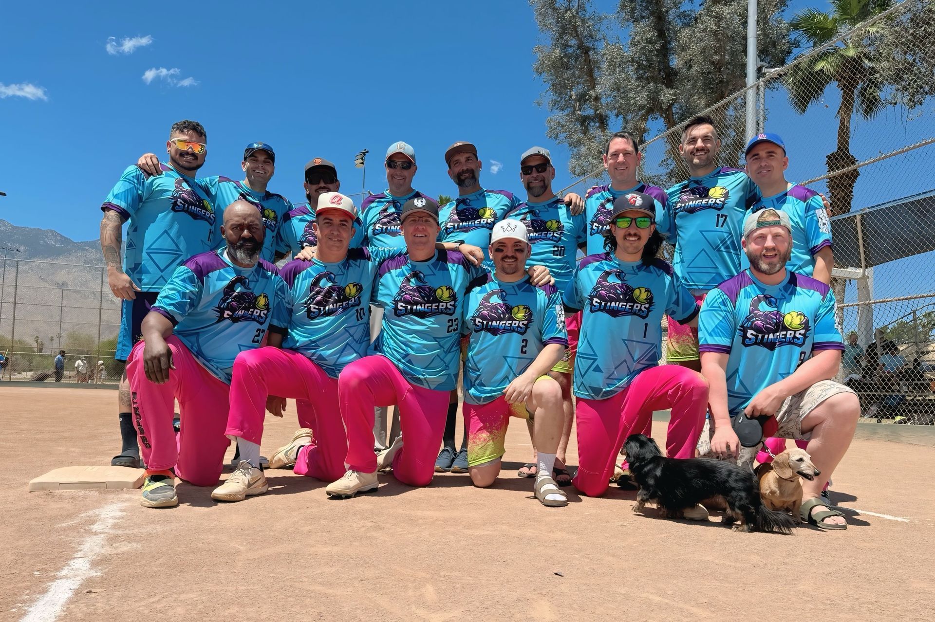 Group of cyclists in blue jerseys and pink shorts posing outdoors under a bright blue sky