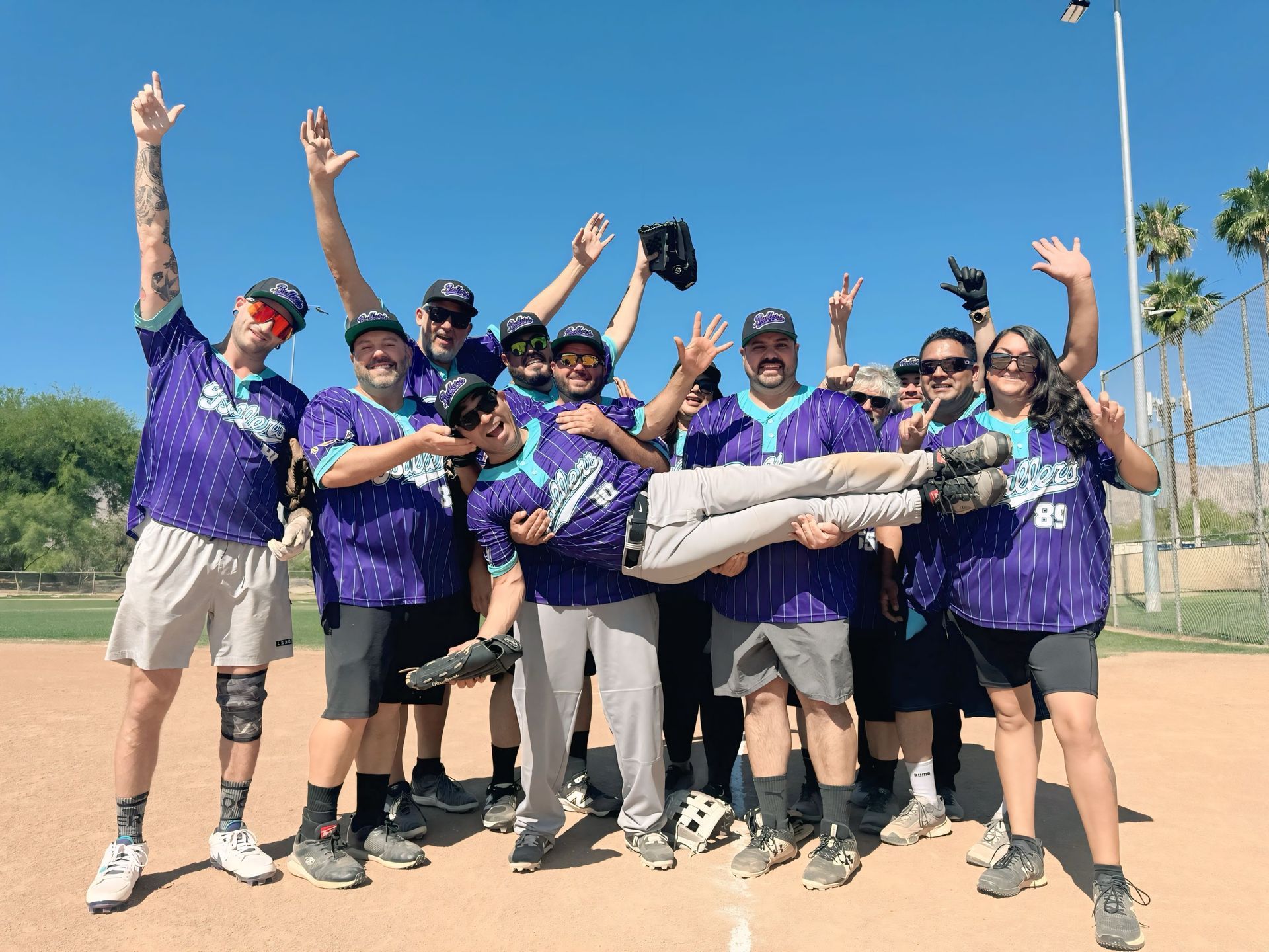 Group of people in purple shirts cheering outdoors, lifting a person horizontally under a bright blue sky