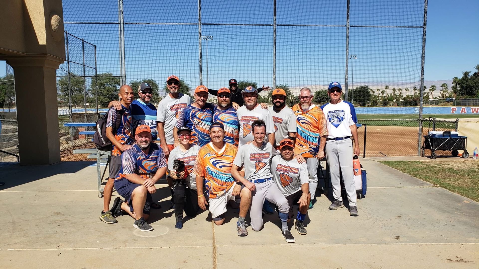 Group of baseball players posing for a team photo on a sunny field, with a blue chain-link fence behind them