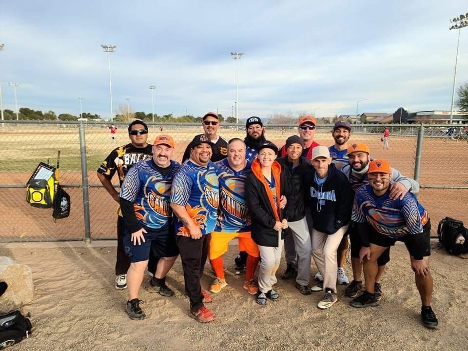 Softball team posing on a dirt field, wearing blue jerseys and orange shorts, with bats and gloves.
