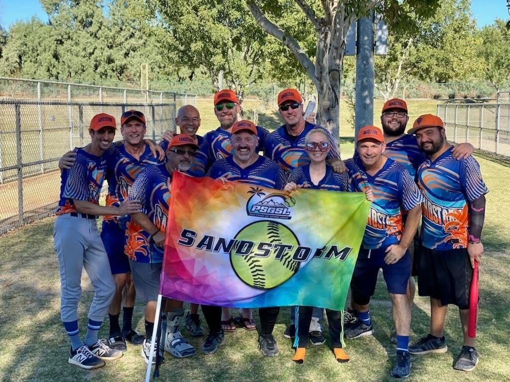 Group of baseball players in blue uniforms holding a colorful team banner outdoors on a field.