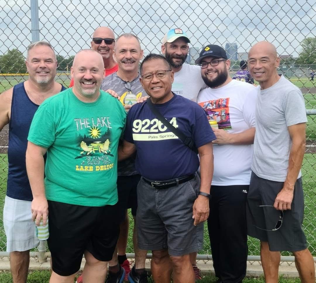 Seven men smiling and posing outdoors in front of a chain-link fence, wearing casual T-shirts.
