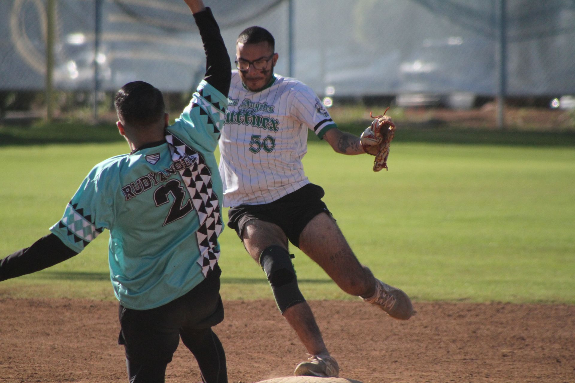 Two athletes in teal and white jerseys collide while reaching for a ball on a dirt field.